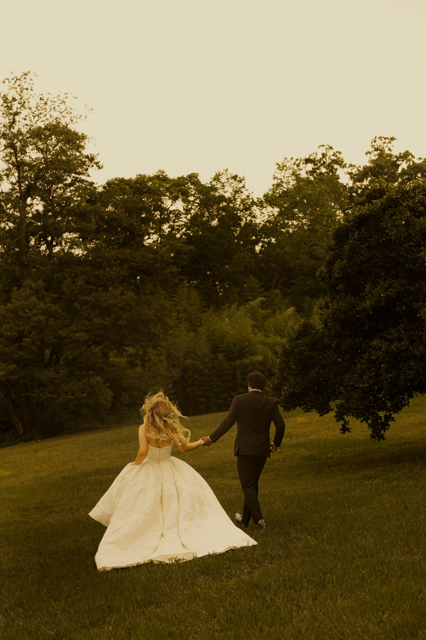 A bride and groom hold hands walking across a lush green lawn at sunset, surrounded by trees.