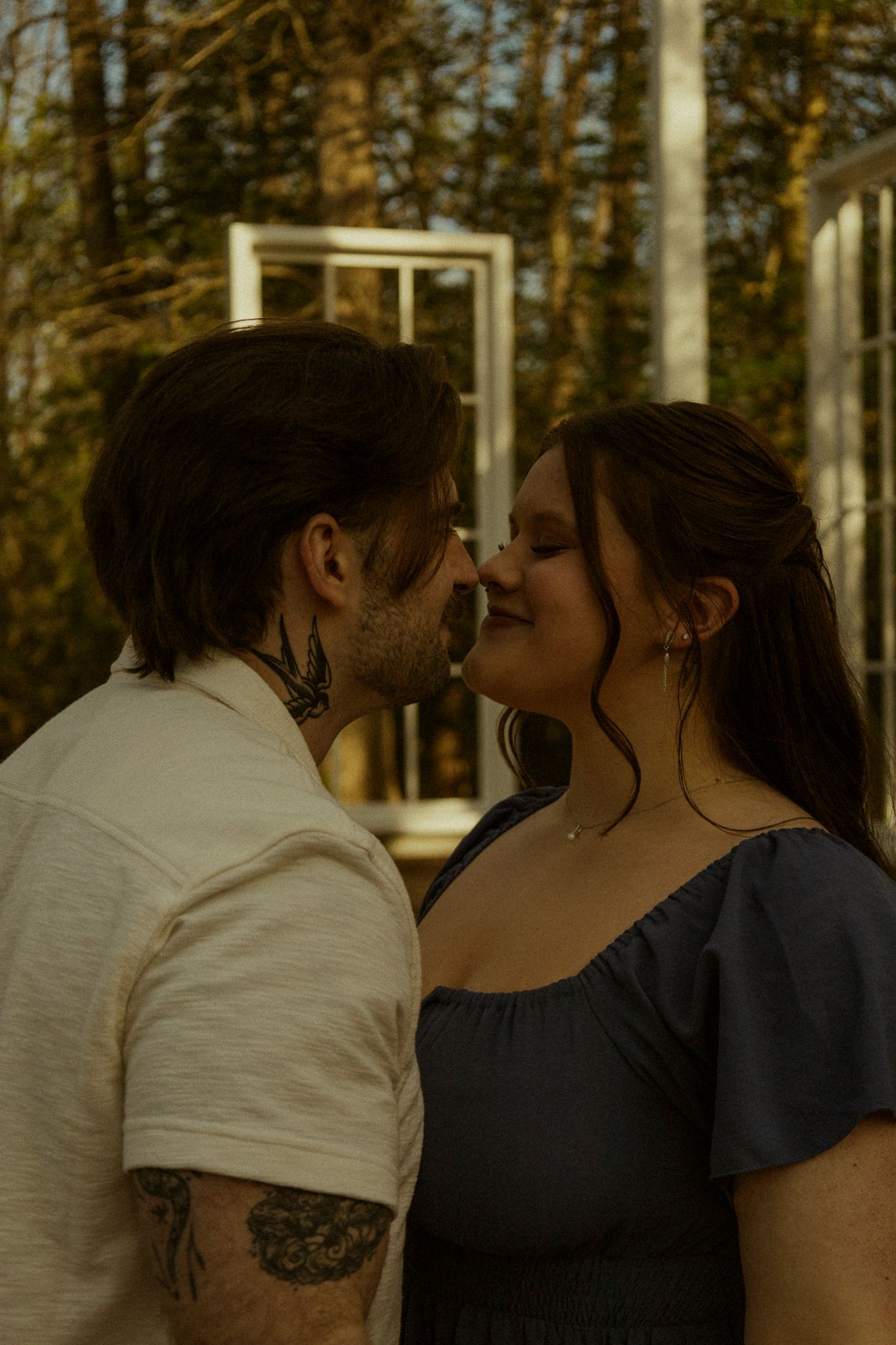 Tattooed couple sharing a tender kiss outdoors with white window frames and forest in background.