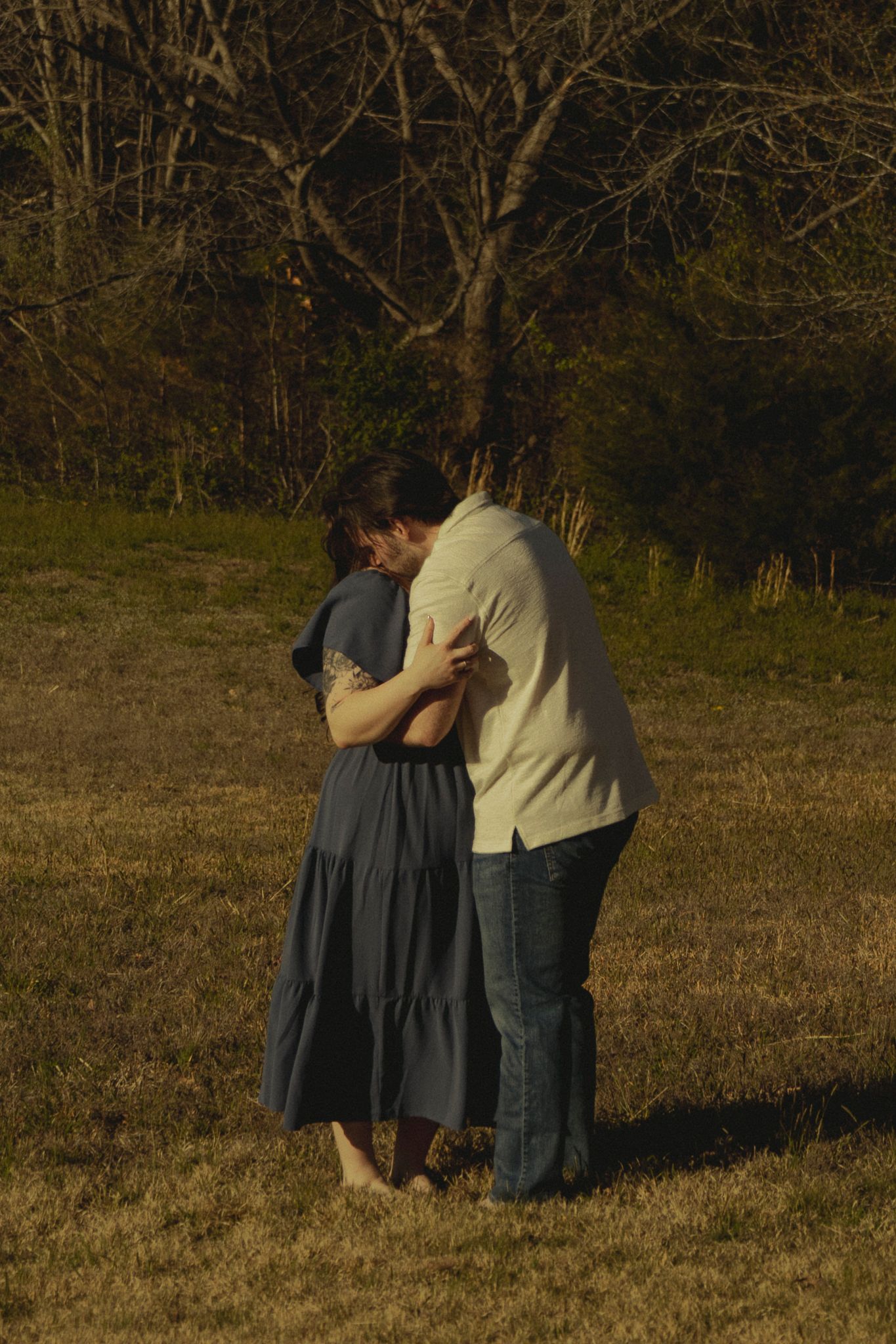 A couple shares a tender embrace in an open field surrounded by trees during golden hour, the woman in a flowing gray dress.