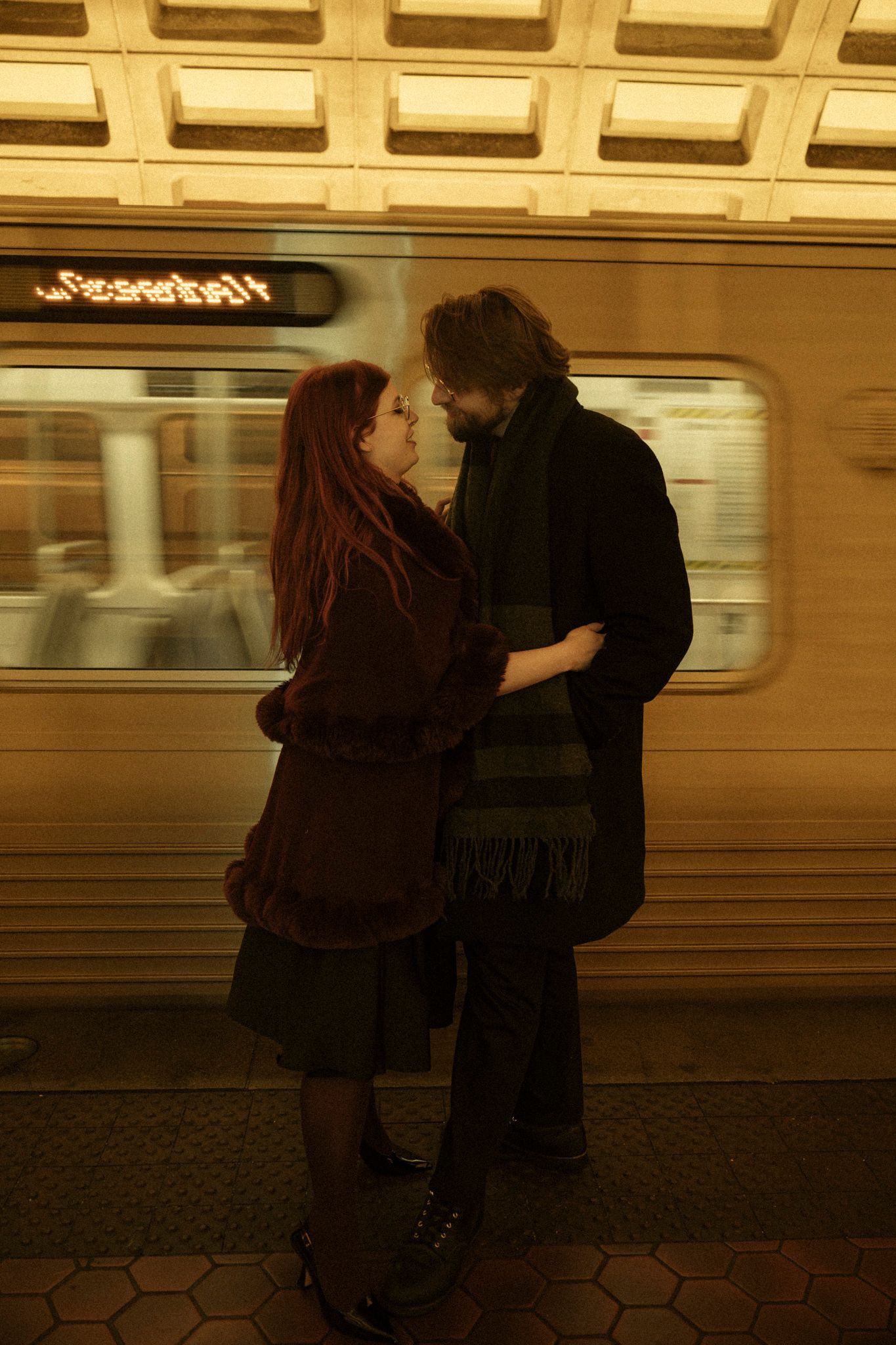 A couple shares an intimate moment on a subway platform with a train car visible in the background.