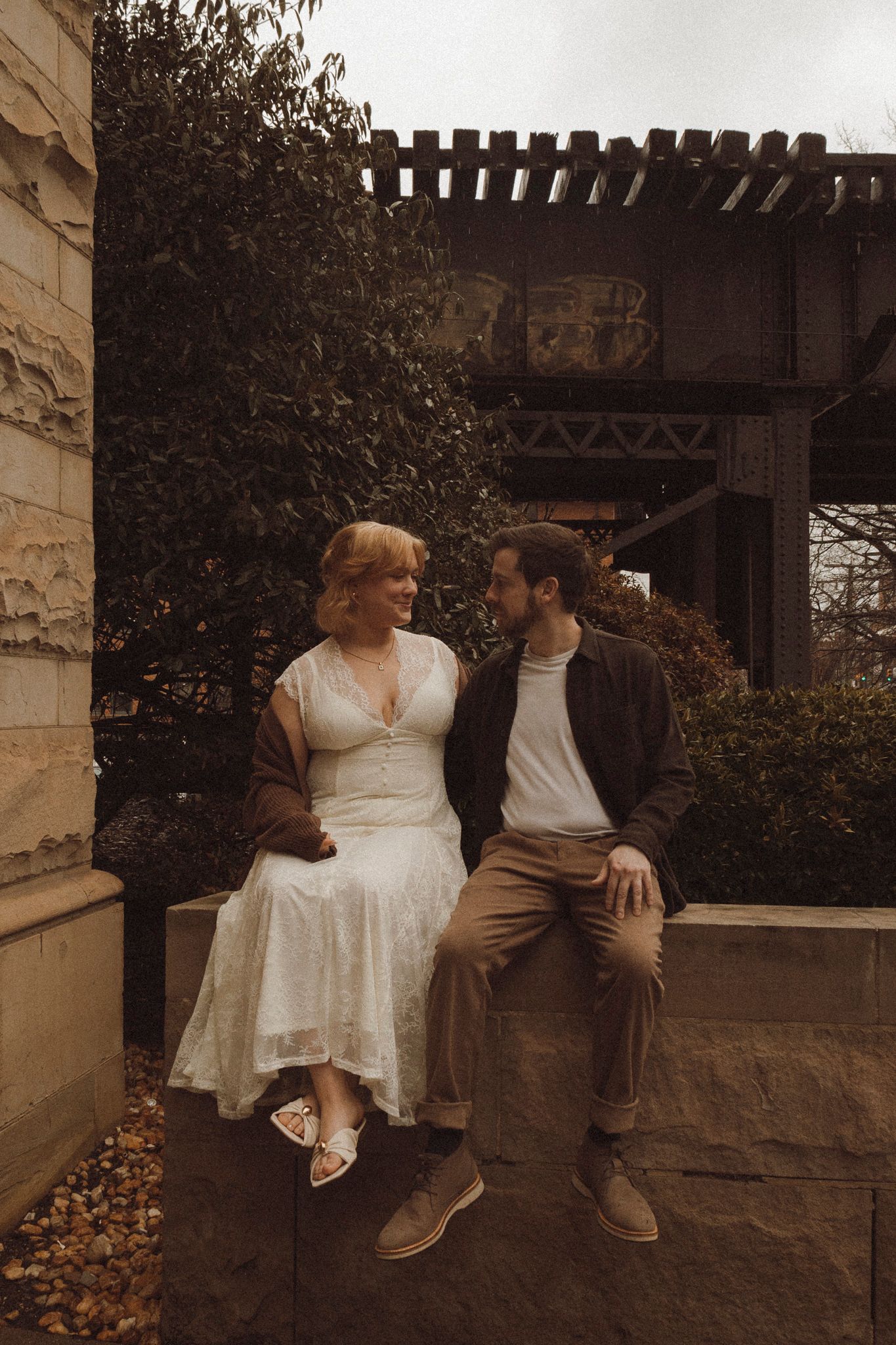 A romantic couple sitting on a stone wall under a wooden pergola in sepia tones.