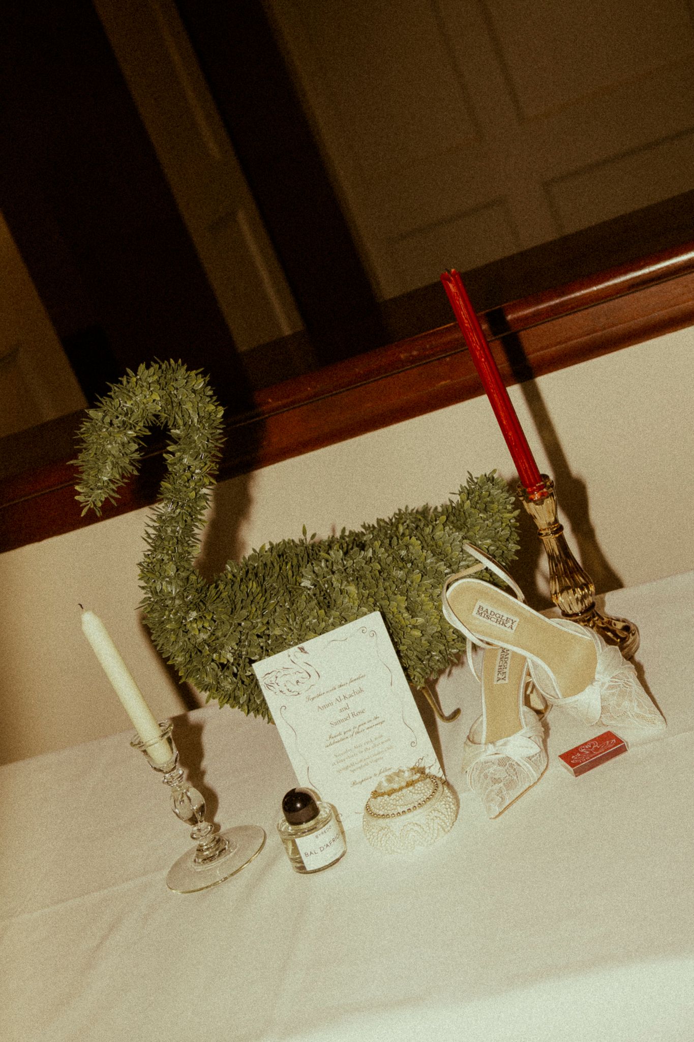 Wedding flat lay with green moss bag, bridal heels, candles, perfume, ring, and invitation on white table.