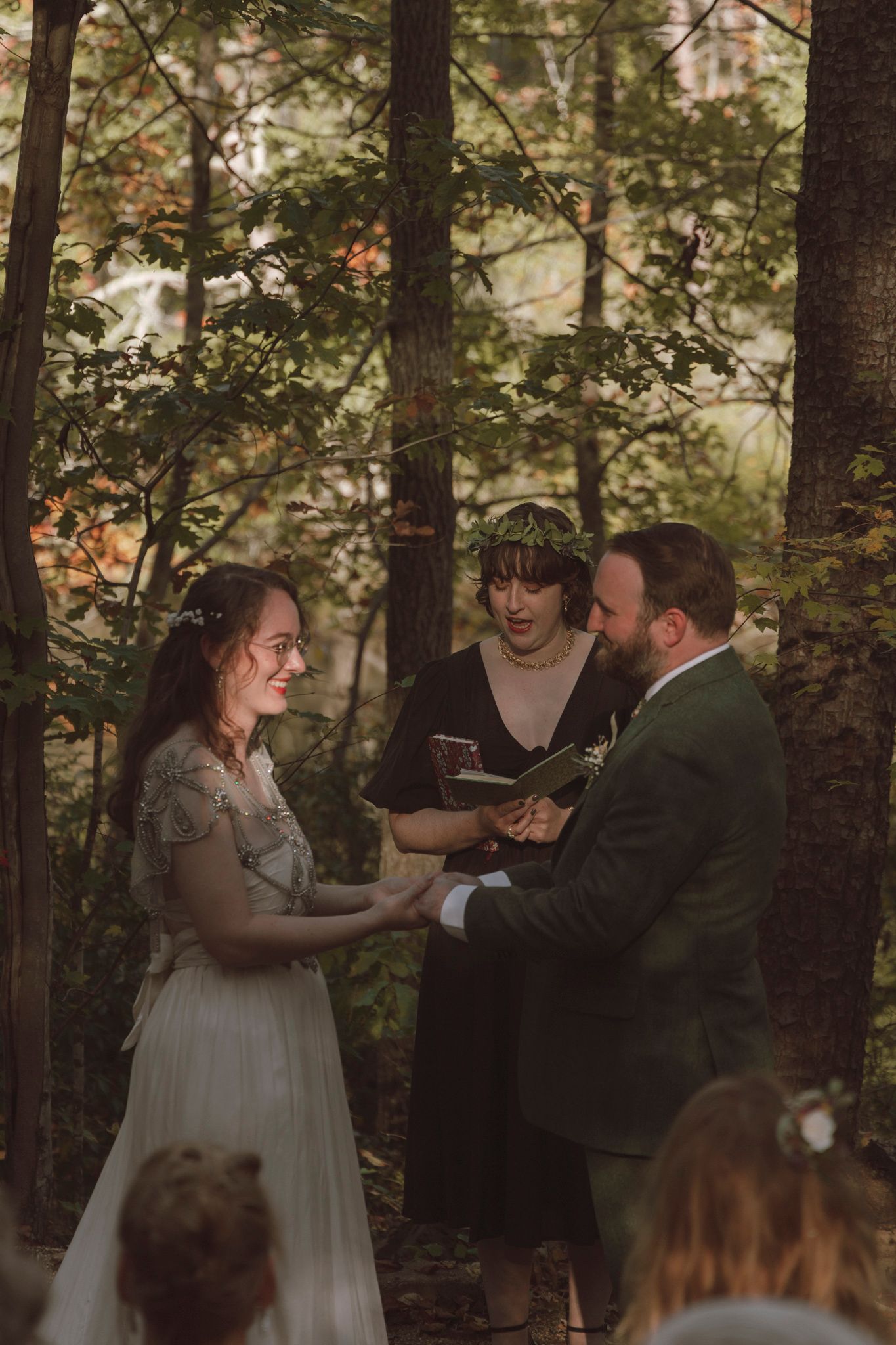 A wedding ceremony takes place in a forest setting with guests gathered among tall trees at sunset.