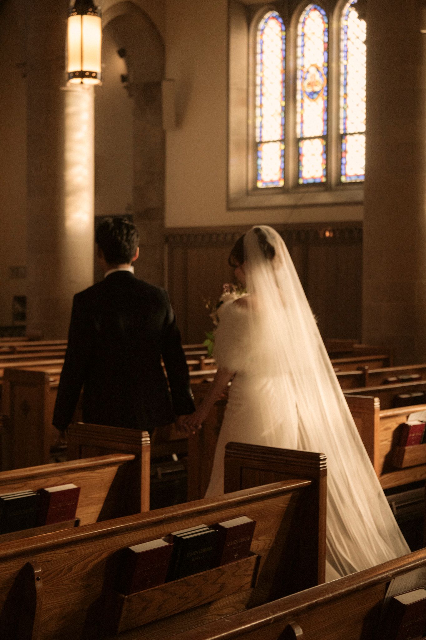 Bride in white gown and veil holds groom's hand while walking toward altar in a warmly lit church.