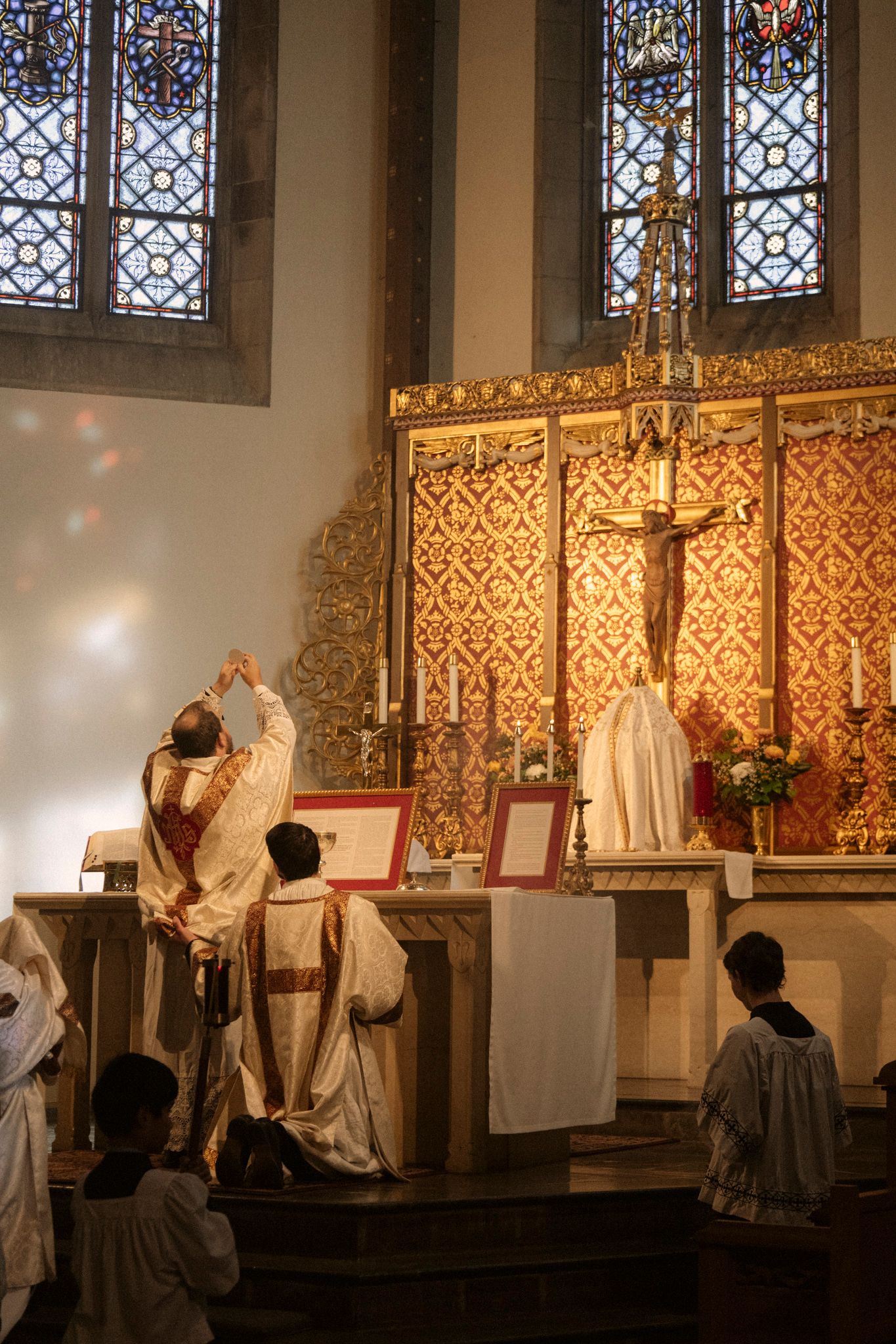 A priest elevates the Eucharist during Catholic Mass at an ornate golden altar with stained glass windows.