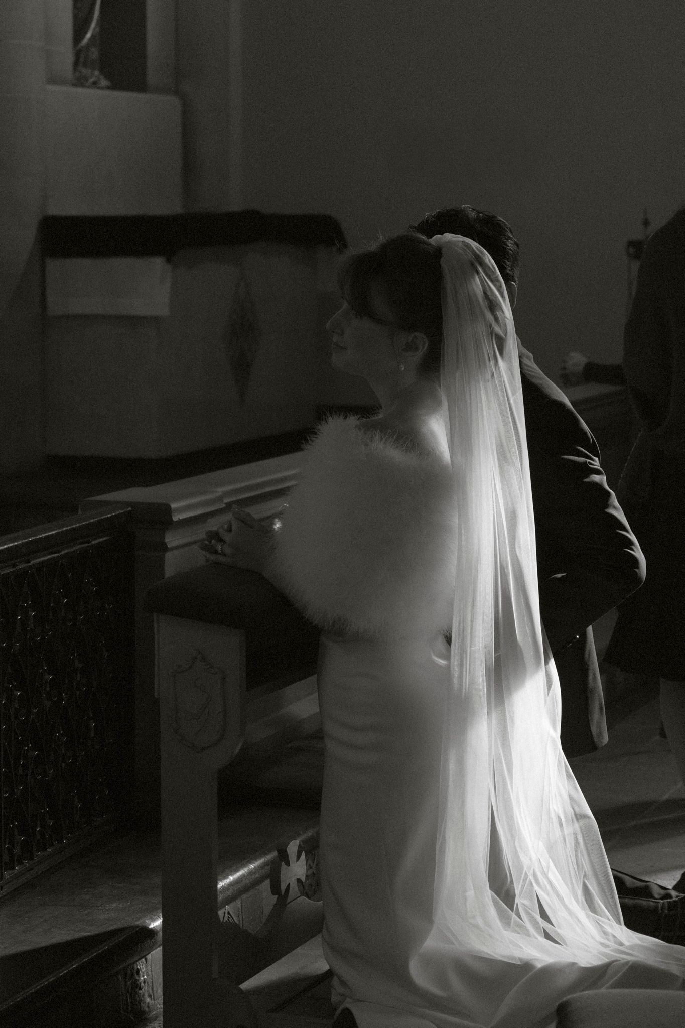 Black and white photo of bride in veil and groom kneeling at church altar.