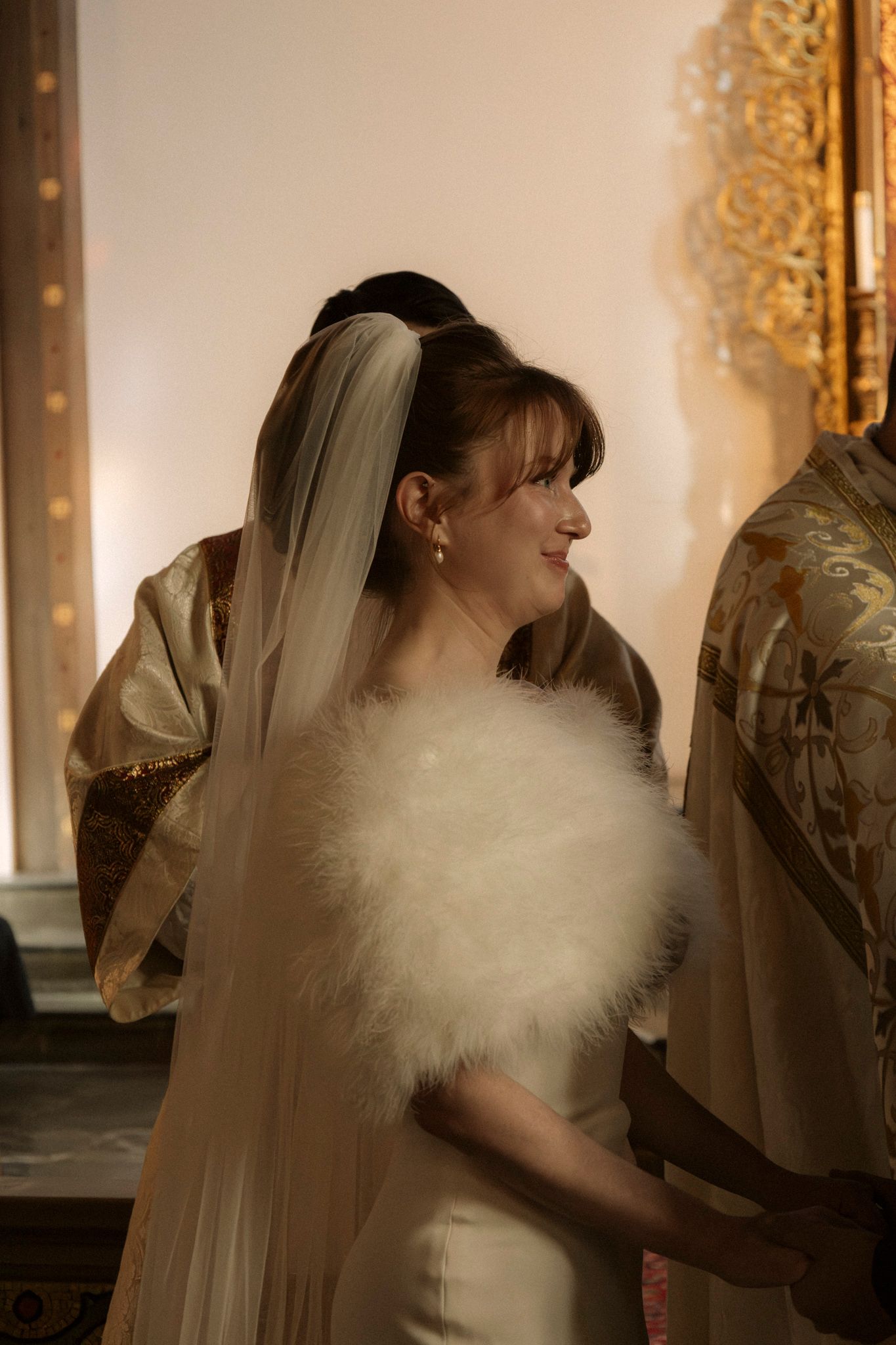 Bride in white satin gown with feather wrap and veil smiles during wedding ceremony.