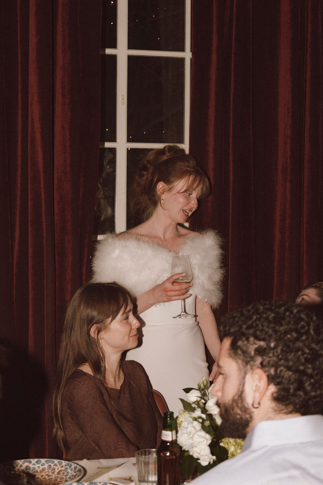 Bride in white strapless gown with fur shawl giving toast at wedding reception dinner.