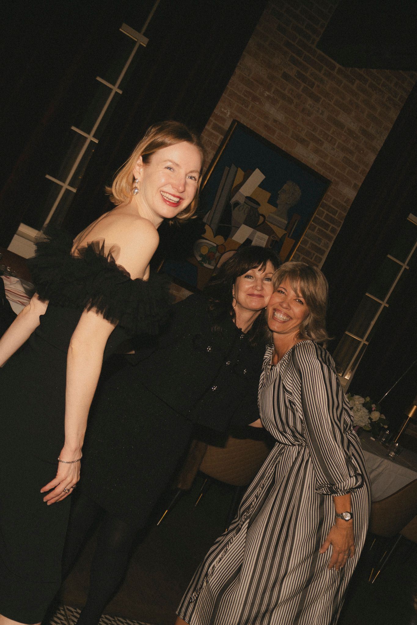 Three women pose and smile together at an elegant wedding reception venue with brick walls.