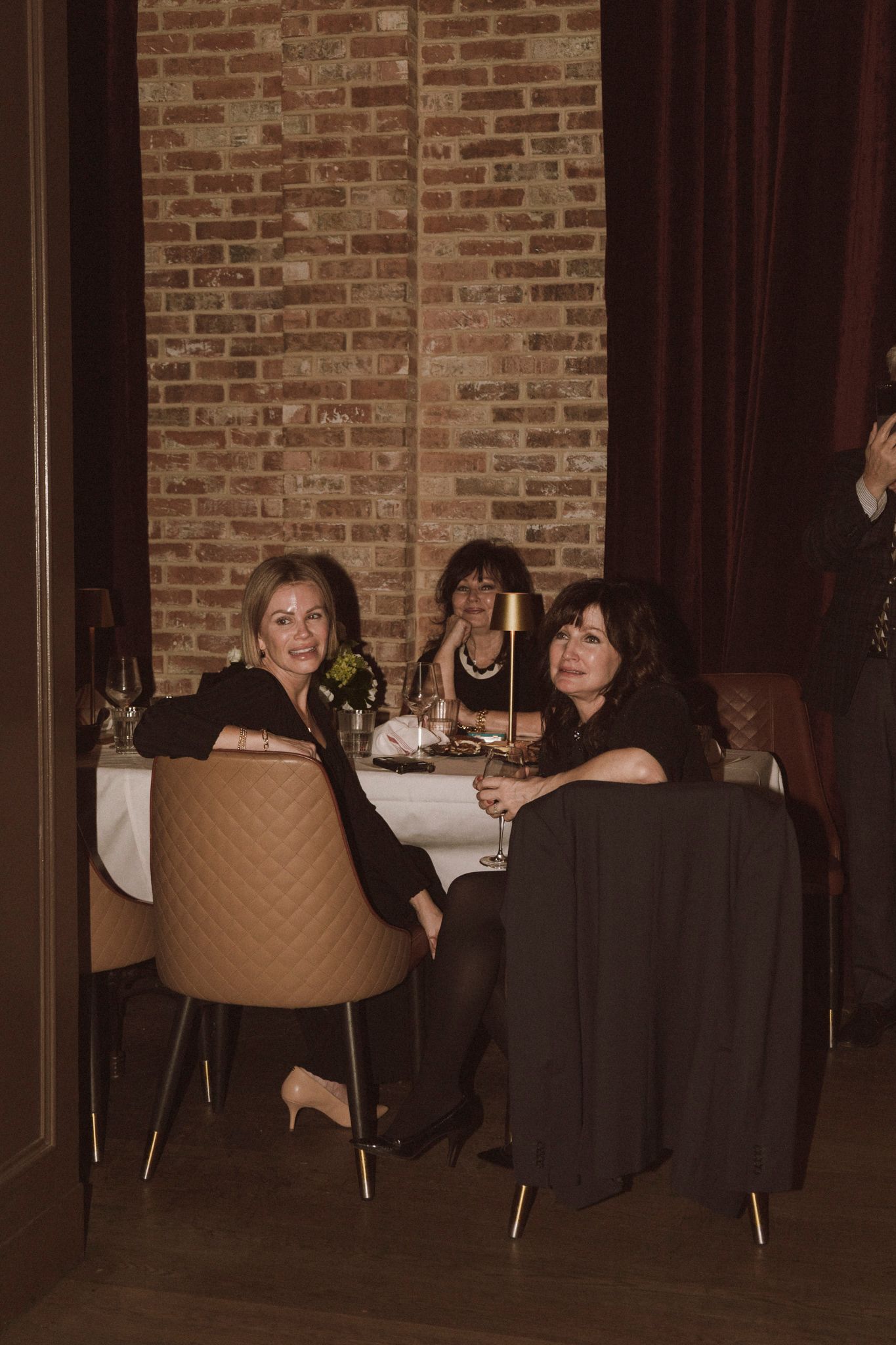 Three women in elegant black attire dining at a restaurant table with brick wall backdrop and gold candlelight.