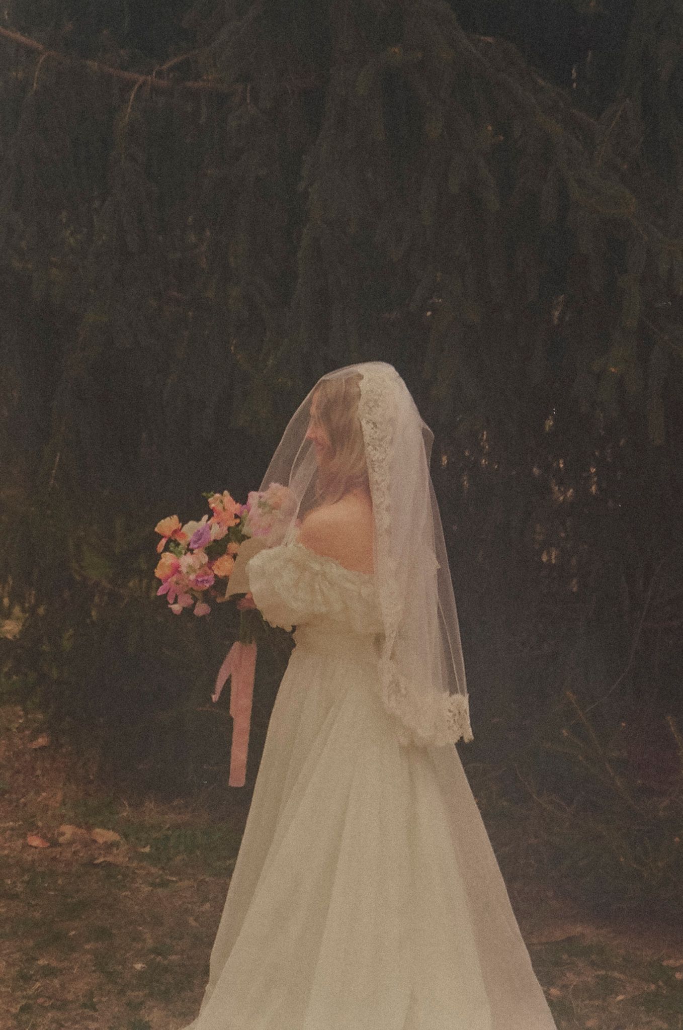 A bride in a flowing white wedding dress and veil stands in a moody forest setting with pink floral accents.