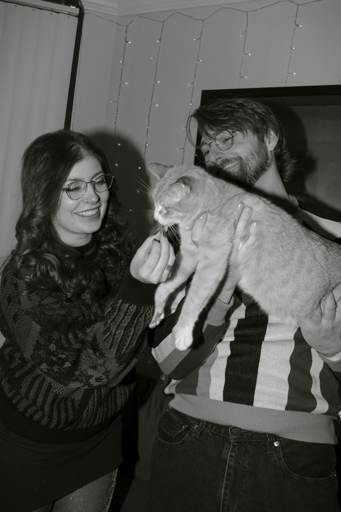 A vintage black and white photograph shows two people in glasses holding and playing with a light-colored cat.