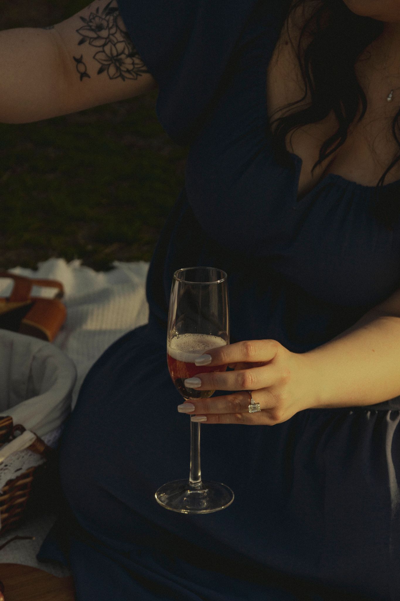 Woman in navy dress holding champagne flute at outdoor picnic, featuring floral arm tattoo and diamond engagement ring.