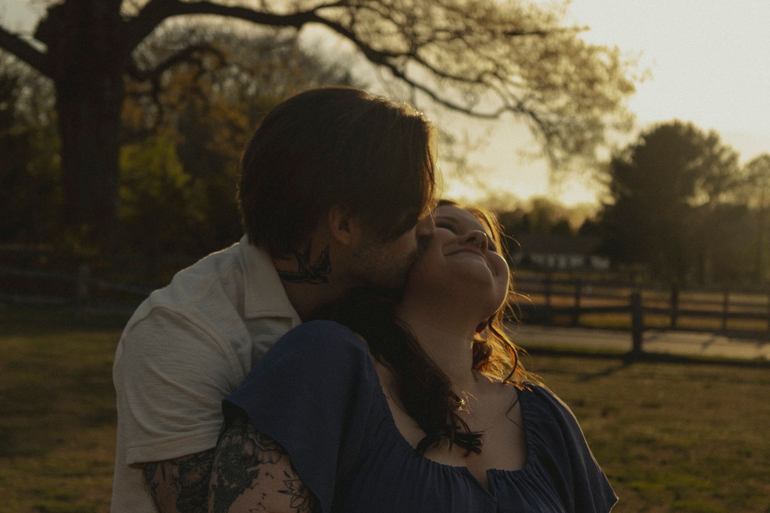 Couple sharing a romantic kiss at golden hour in a park with trees and fence in background.