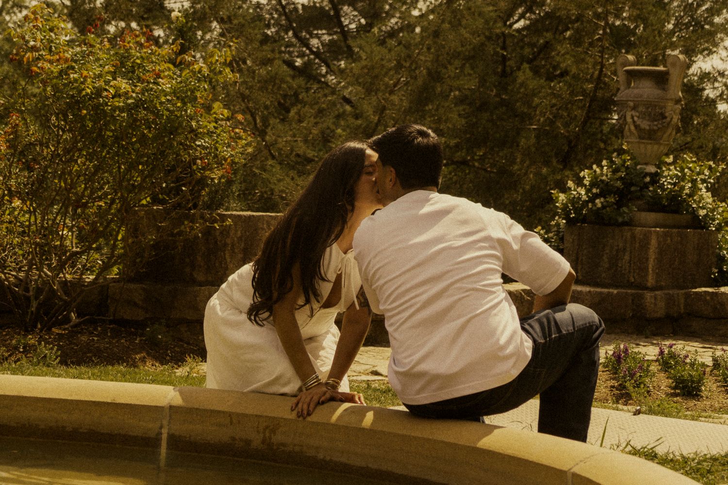 Romantic couple sharing a tender kiss by a garden fountain surrounded by lush green trees.