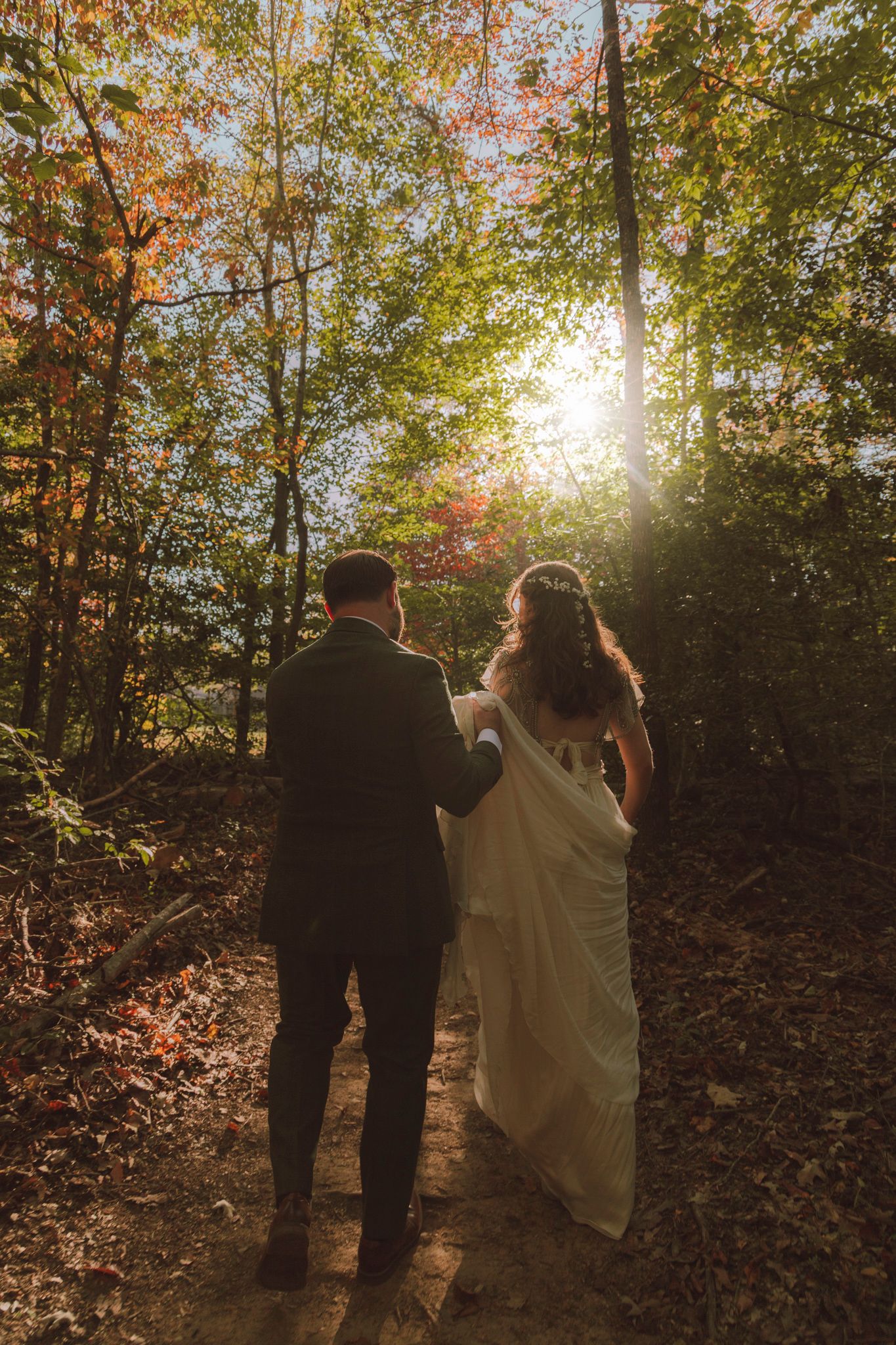 A couple in wedding attire walks along a sunlit forest path surrounded by autumn foliage during golden hour.