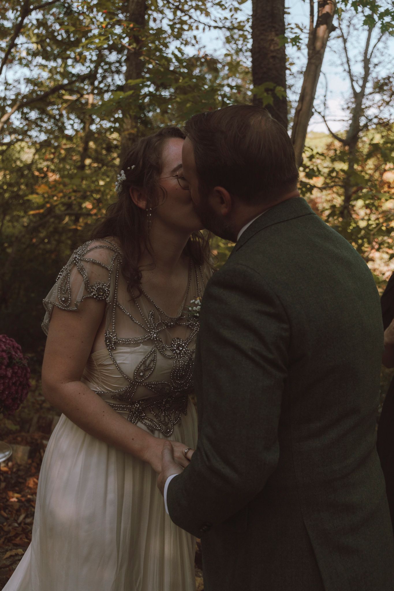 A romantic outdoor autumn wedding scene with a couple sharing a kiss among trees in golden sunlight.