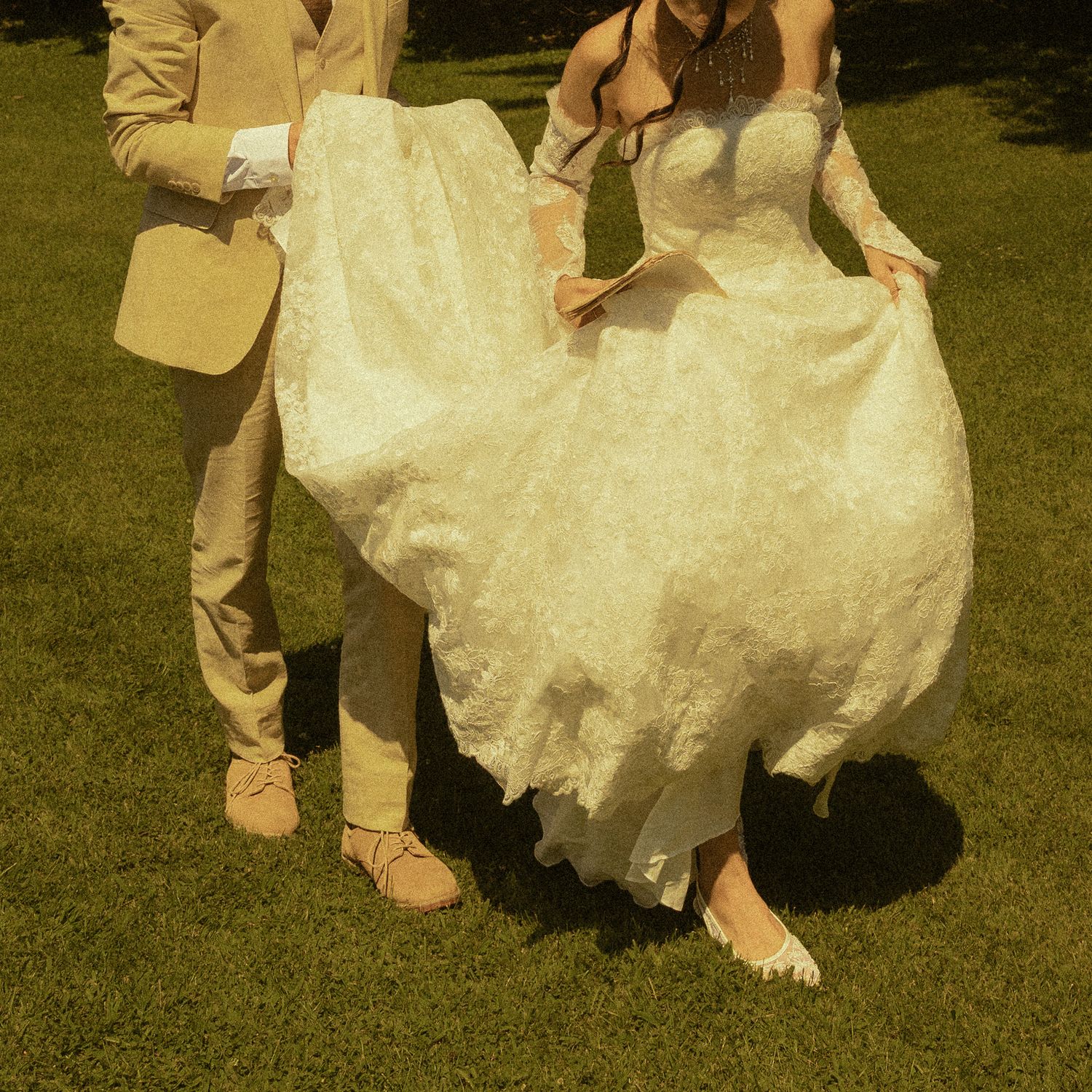 Bride in lace wedding gown and groom in beige suit standing on green grass at outdoor wedding ceremony.