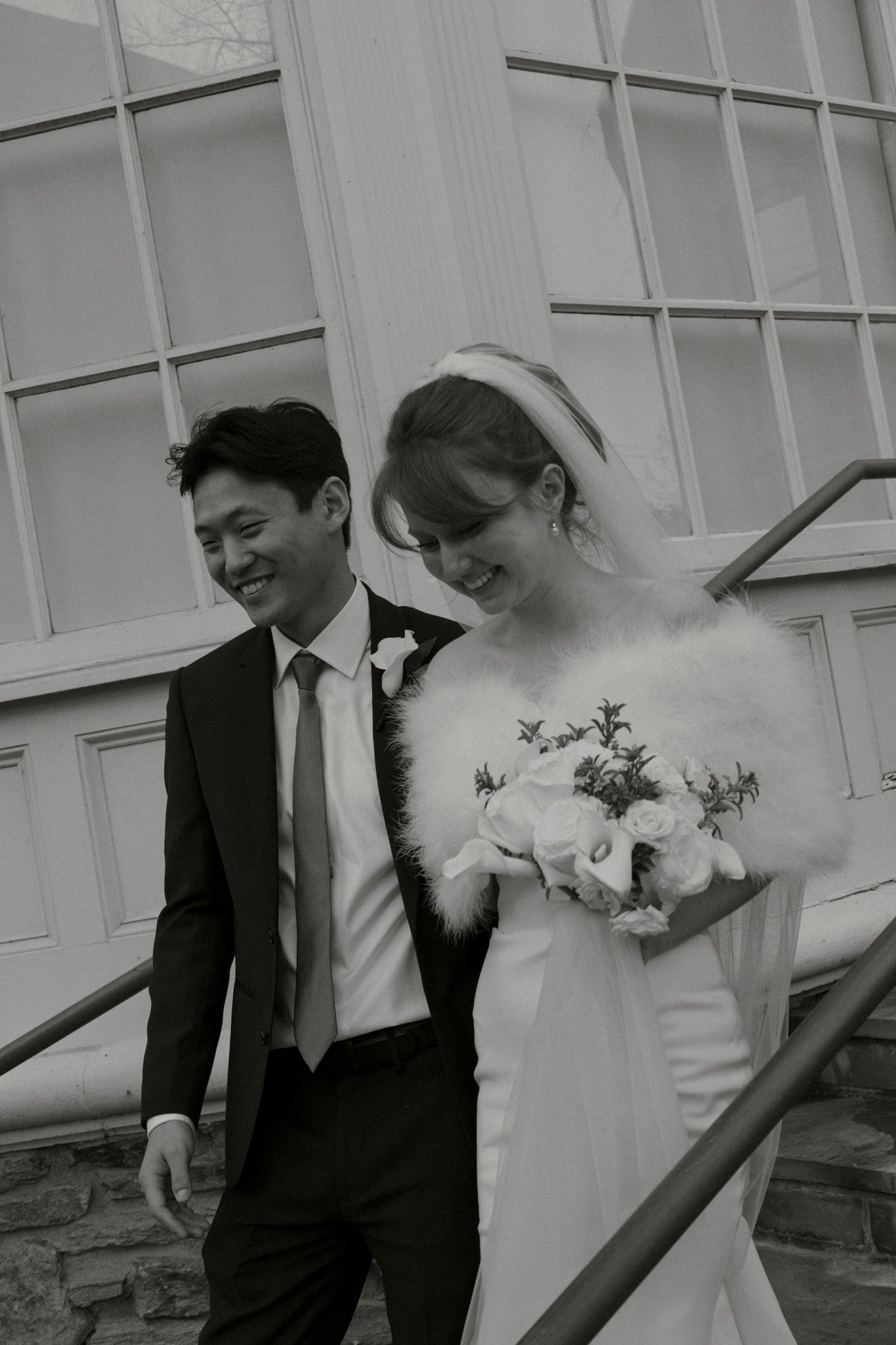 Happy bride and groom walk down outdoor steps in black and white wedding portrait with bouquet.