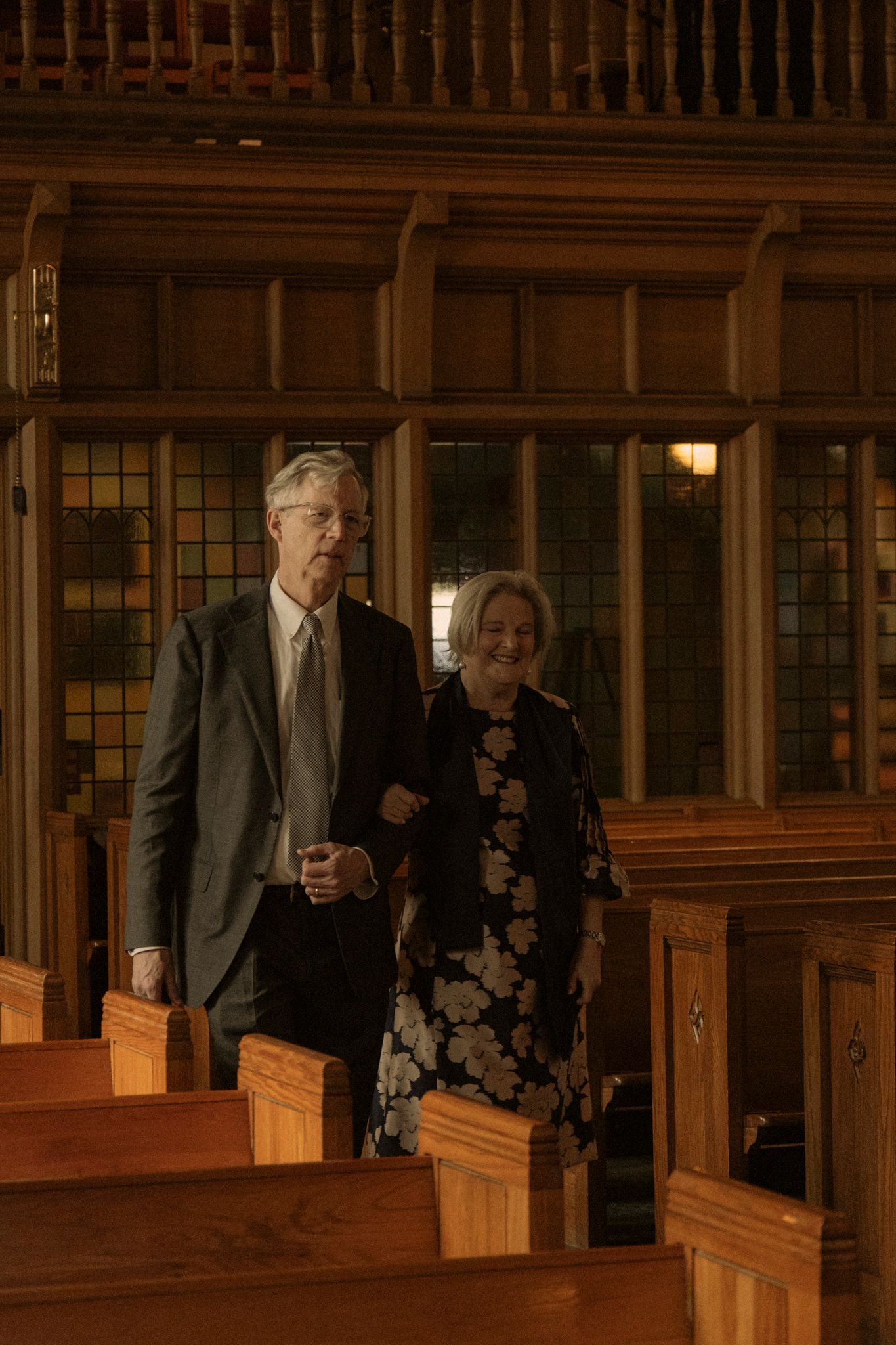 Elderly couple walking together through a warmly lit wooden church interior with stained glass windows.