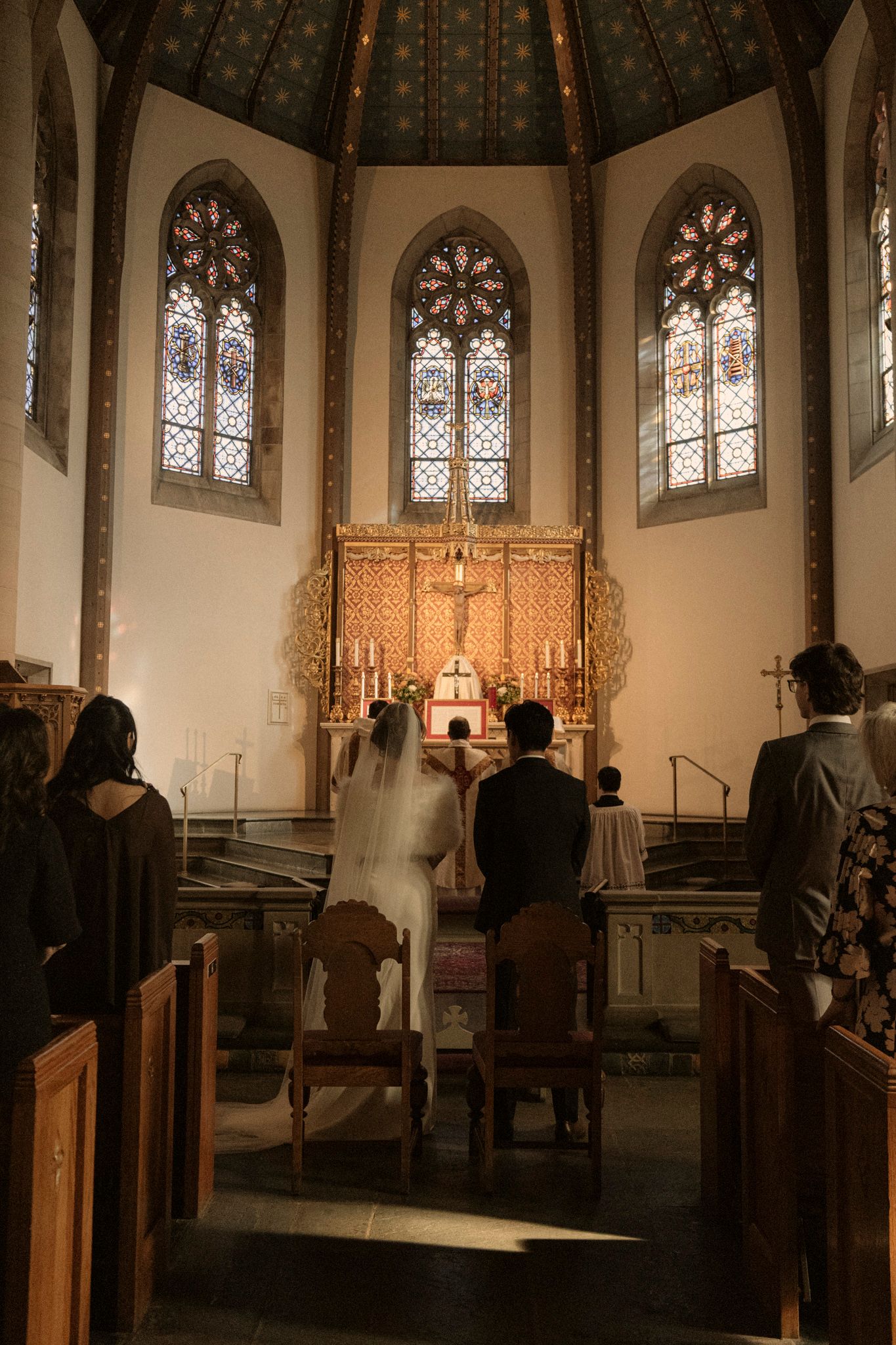 A bride and groom kneel before a golden altar during a church wedding ceremony with stained glass windows.
