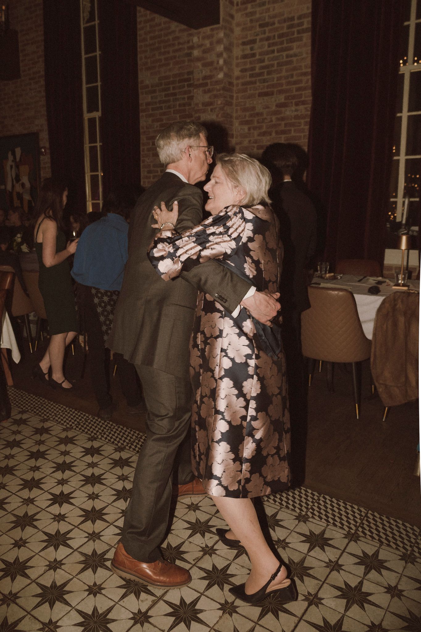 Older couple dancing together joyfully on patterned tile floor at wedding reception.