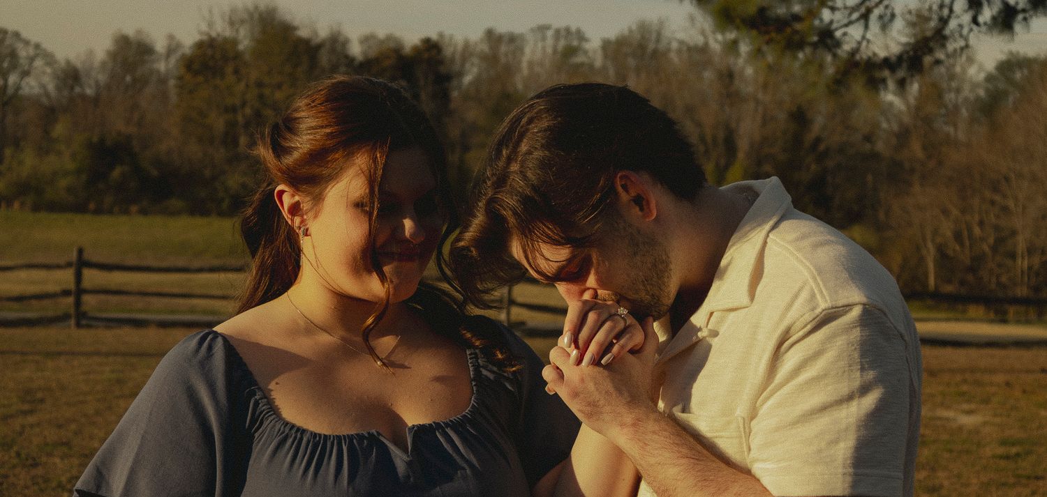 A couple shares a tender moment outdoors, holding hands near a wooden fence in a warm, golden-lit setting.