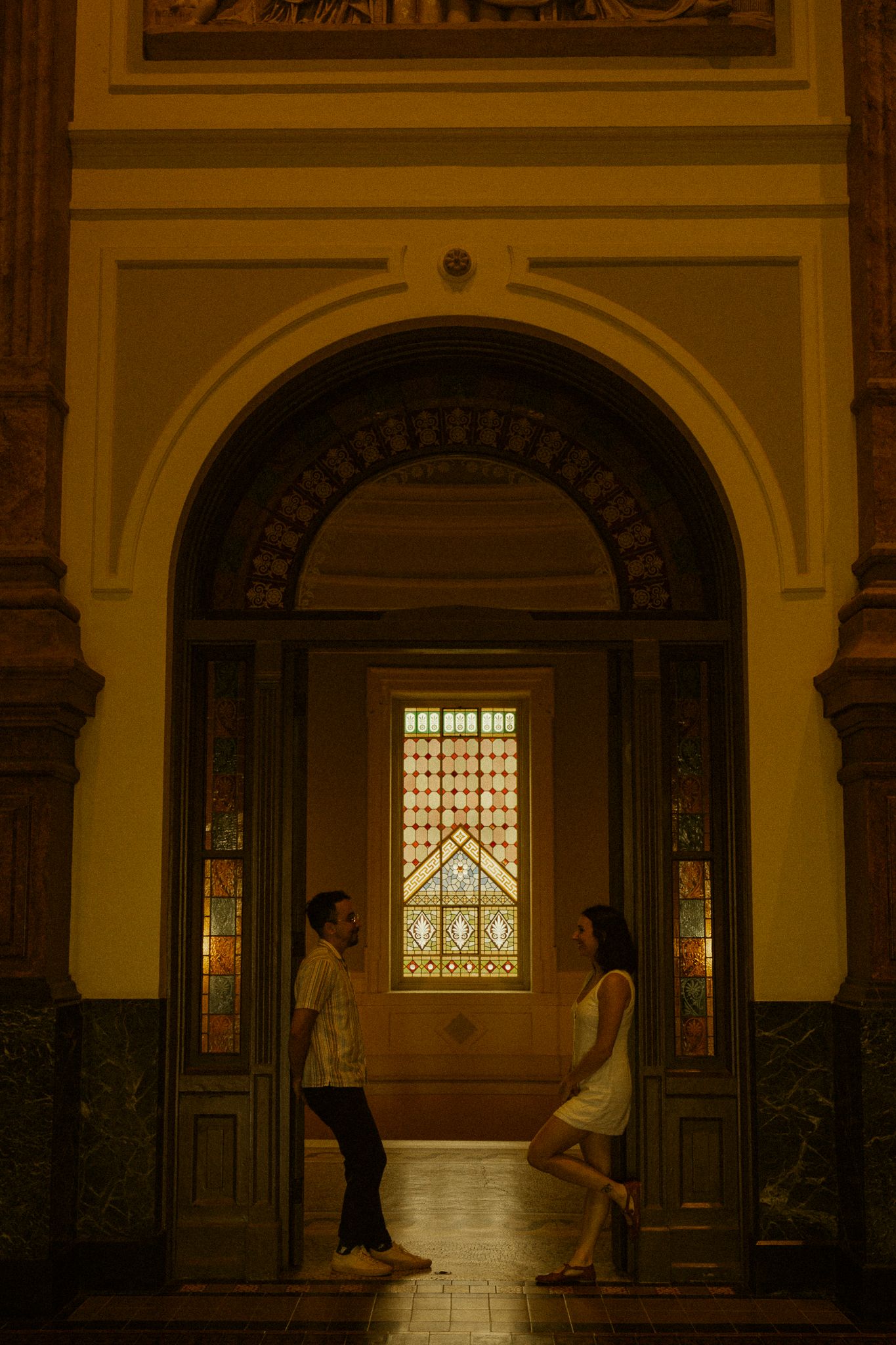 Couple facing each other under ornate arch with stained glass window in elegant historic building.