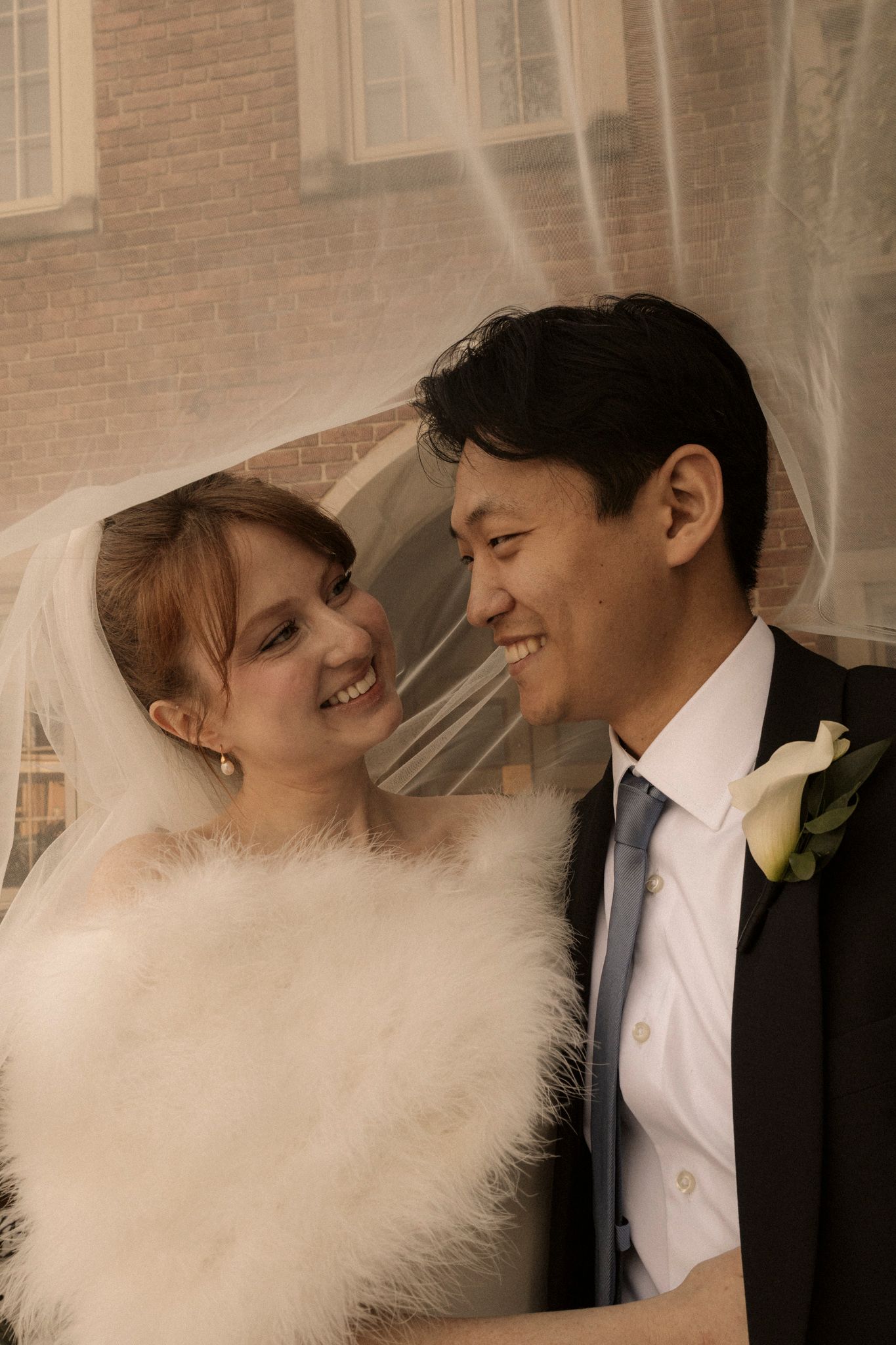 Smiling bride and groom sharing a joyful moment under a flowing wedding veil outside a brick building.