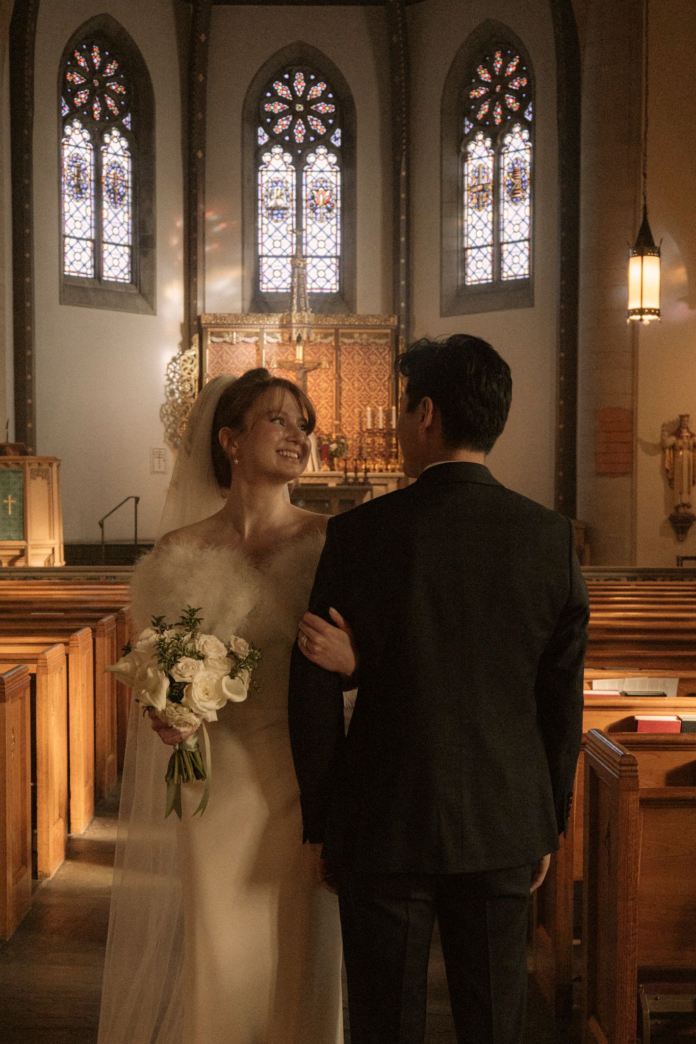 Bride in white gown holding bouquet smiles at groom in dark suit inside a church with stained glass windows.