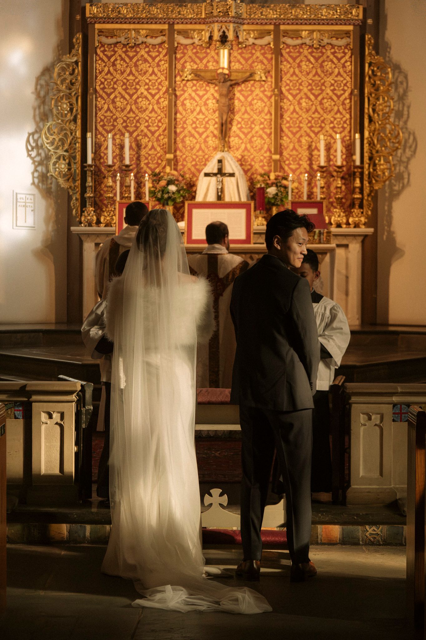 Bride in white veil and groom standing before ornate golden altar with crucifix during church wedding.
