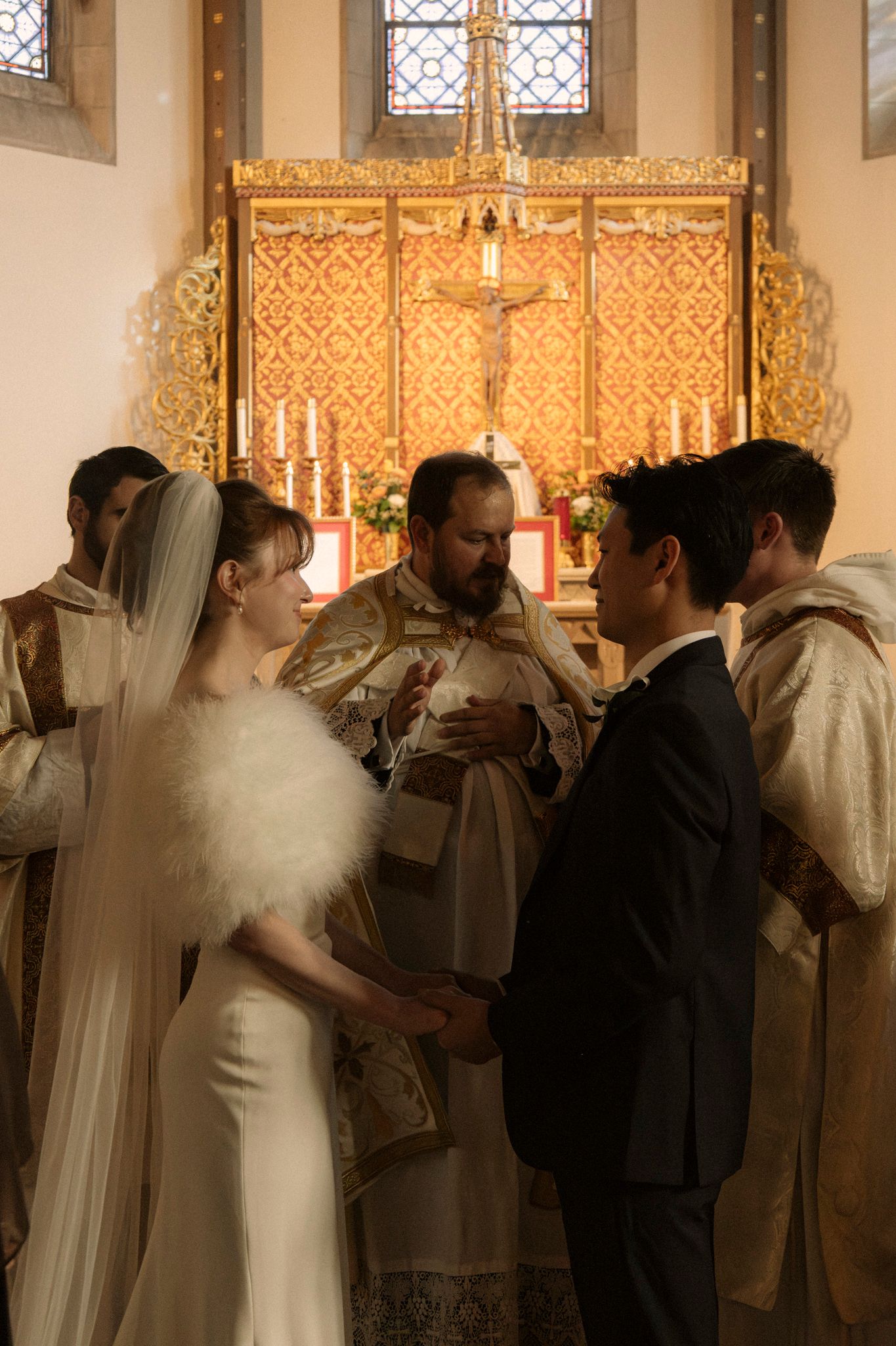 Bride and groom exchange vows before priest at ornate golden altar during traditional Catholic wedding ceremony.