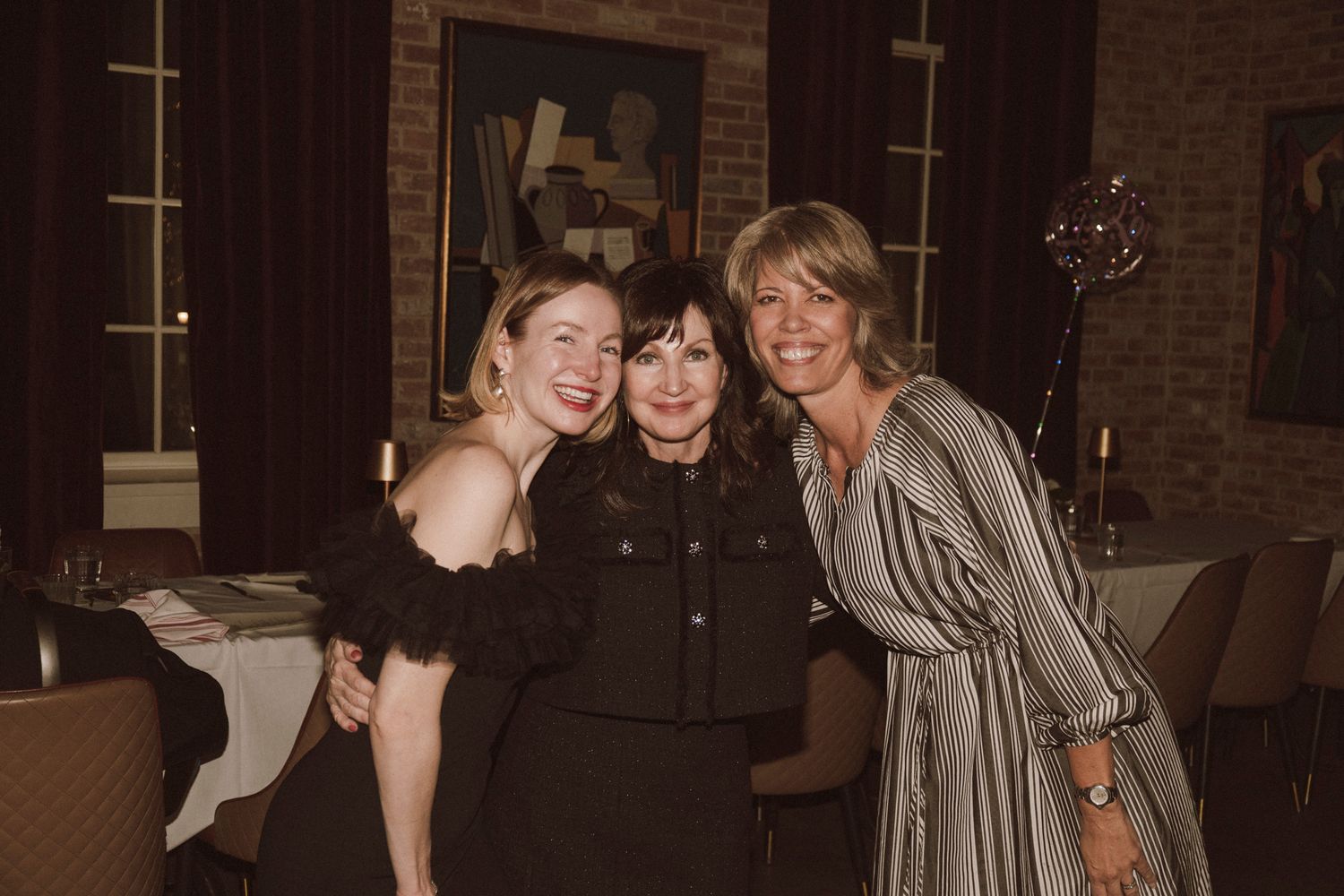 Three women smiling and posing together at an indoor wedding reception in a dimly lit venue.