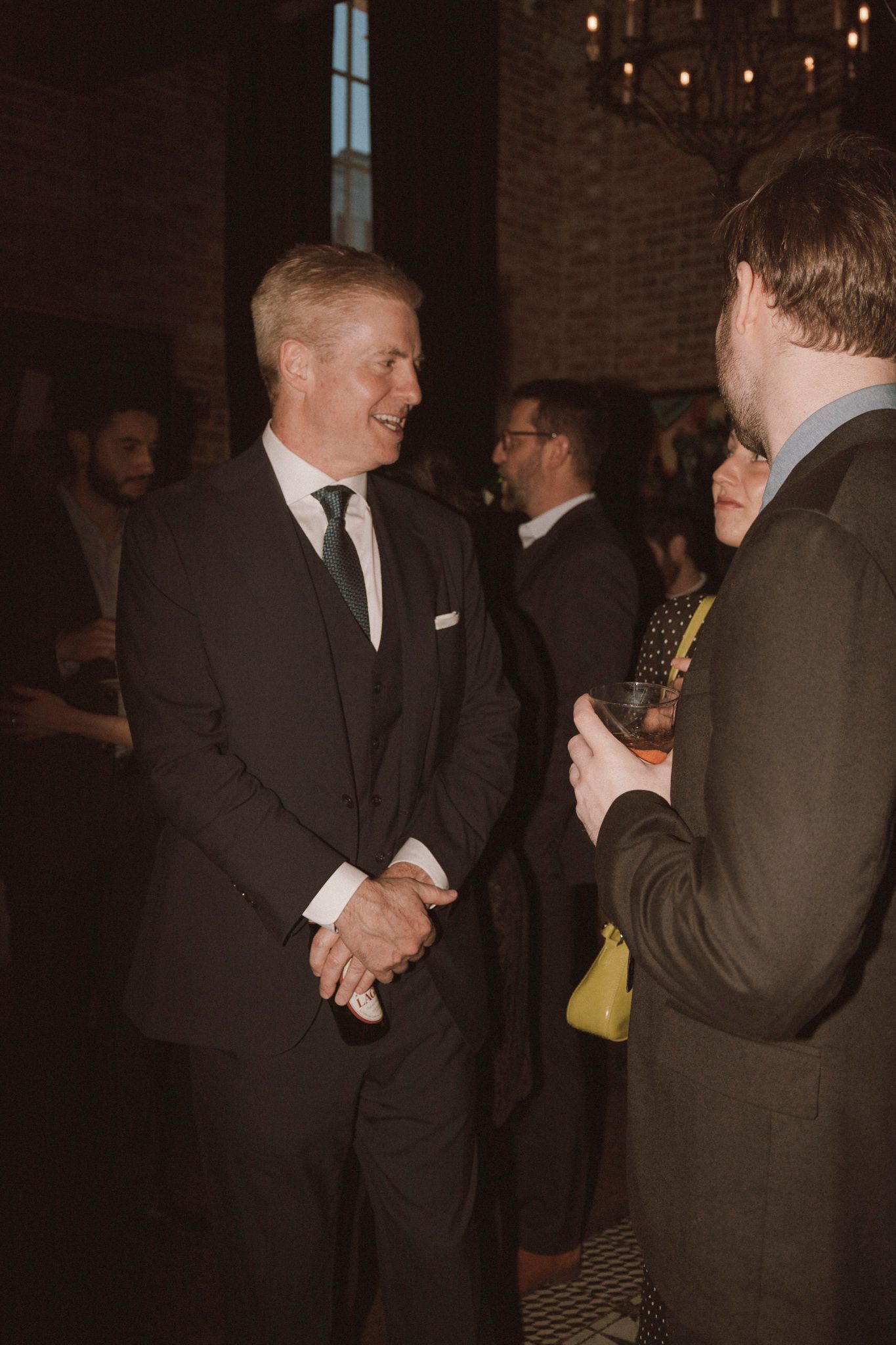 Men in dark suits socializing at an upscale indoor event in a warmly lit brick venue.