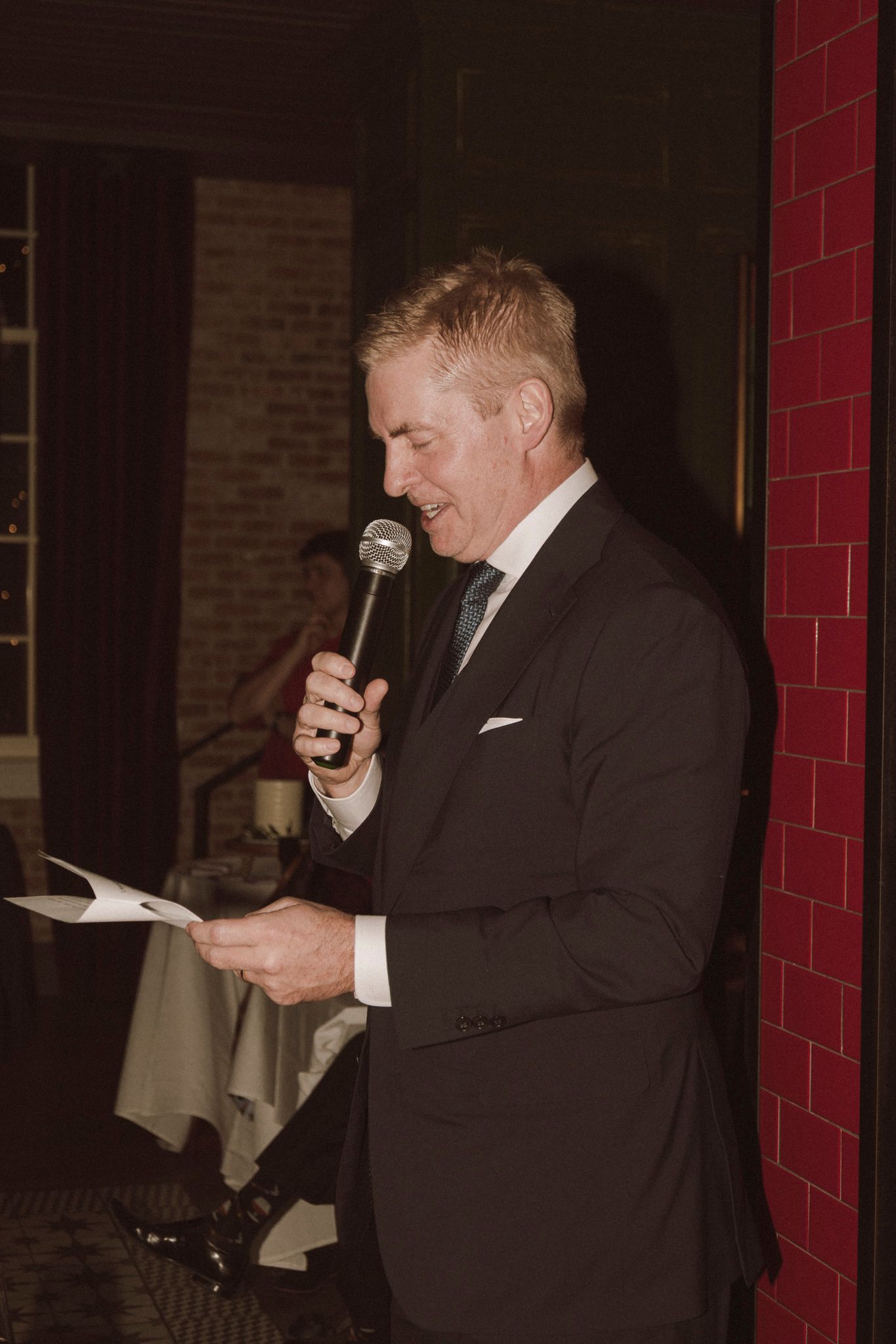 Elegantly dressed man in a dark suit reads a speech into a microphone at a wedding reception.