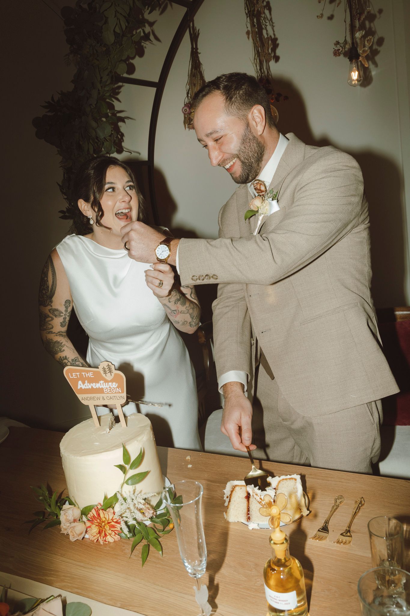 A couple shares a playful moment while cutting their wedding cake at a reception decorated with greenery and flowers.