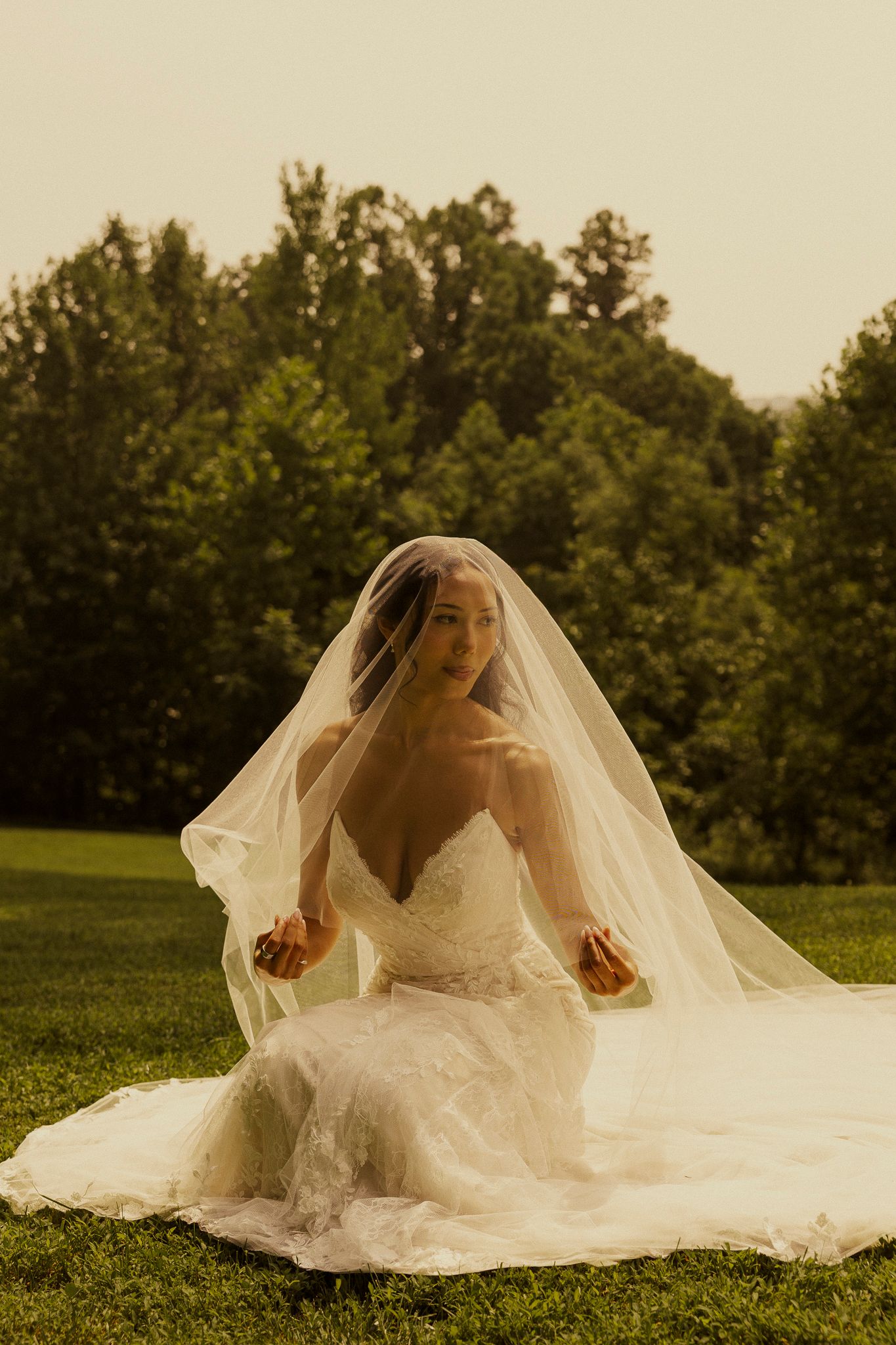 Bride in white lace gown and flowing veil sits on green grass with trees in background during outdoor wedding portrait.