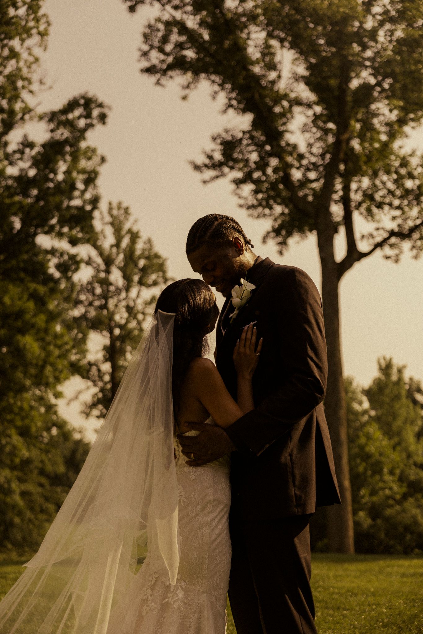 A bride and groom share a romantic kiss outdoors, surrounded by lush green trees during golden hour.