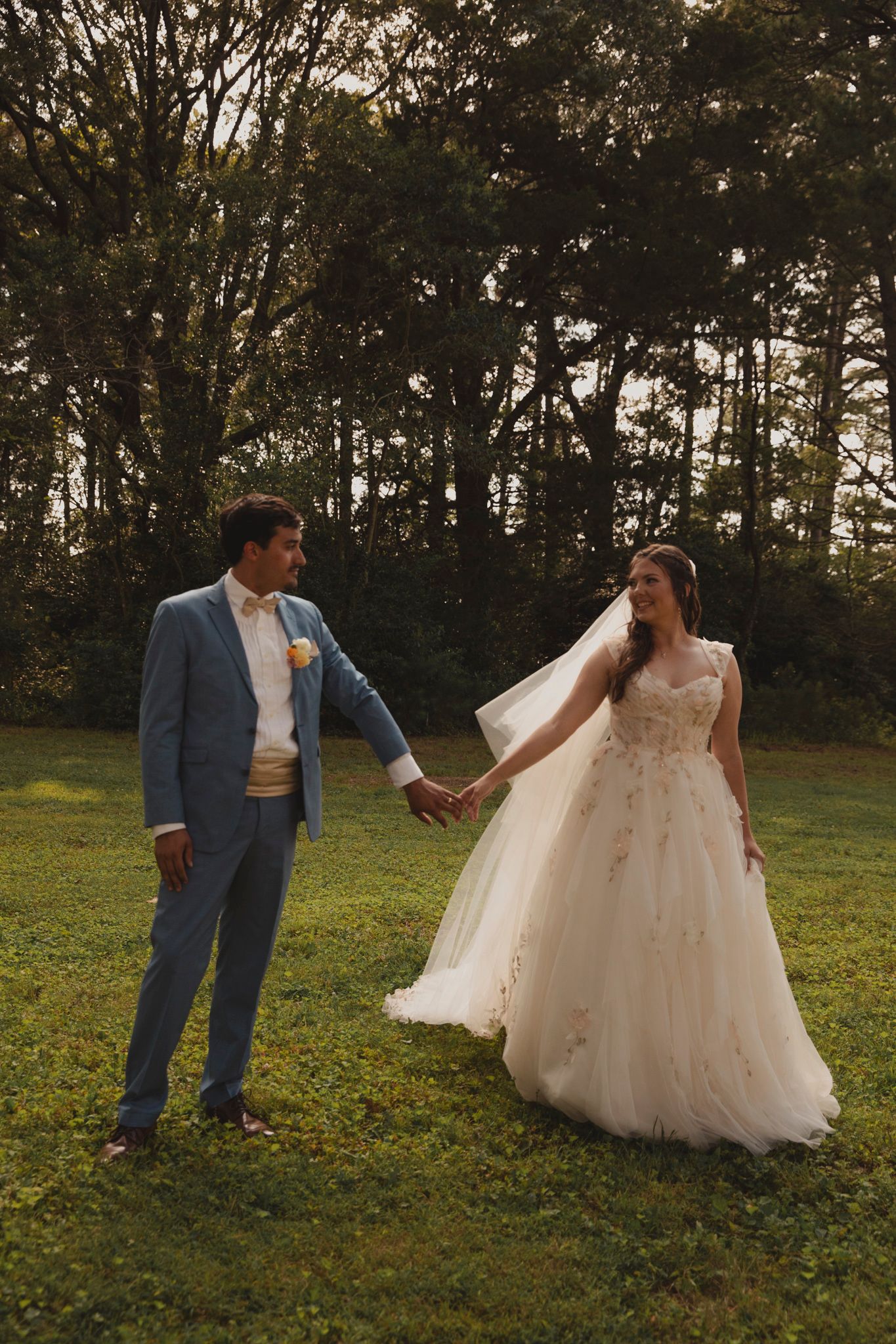 A romantic couple in wedding attire share playful moments on a grassy field at sunset with trees in the background.