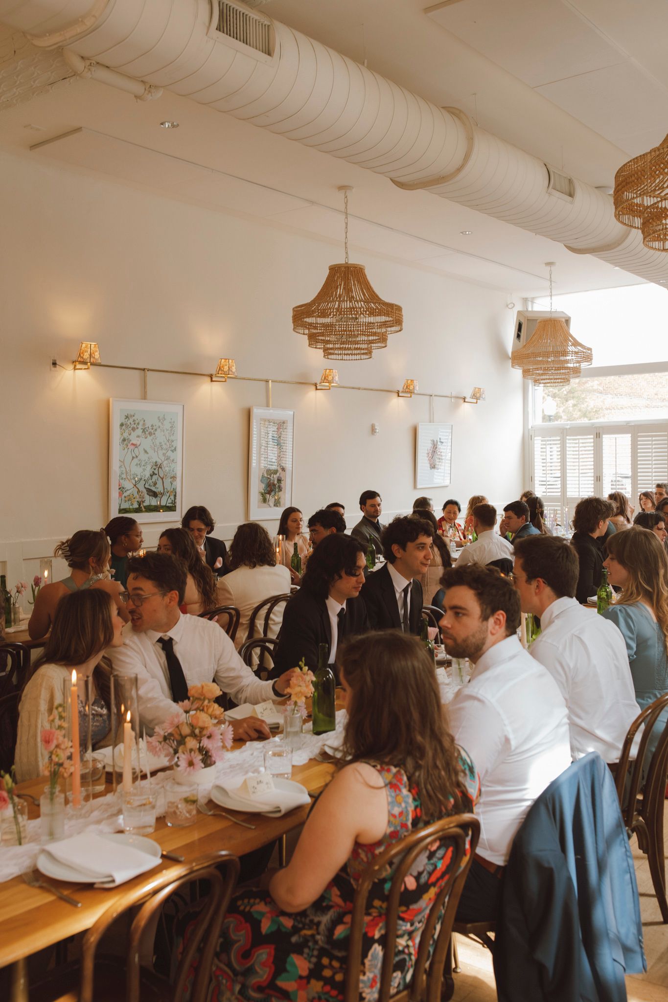 Guests seated at long communal tables in a bright modern restaurant space with white walls and hanging light fixtures.