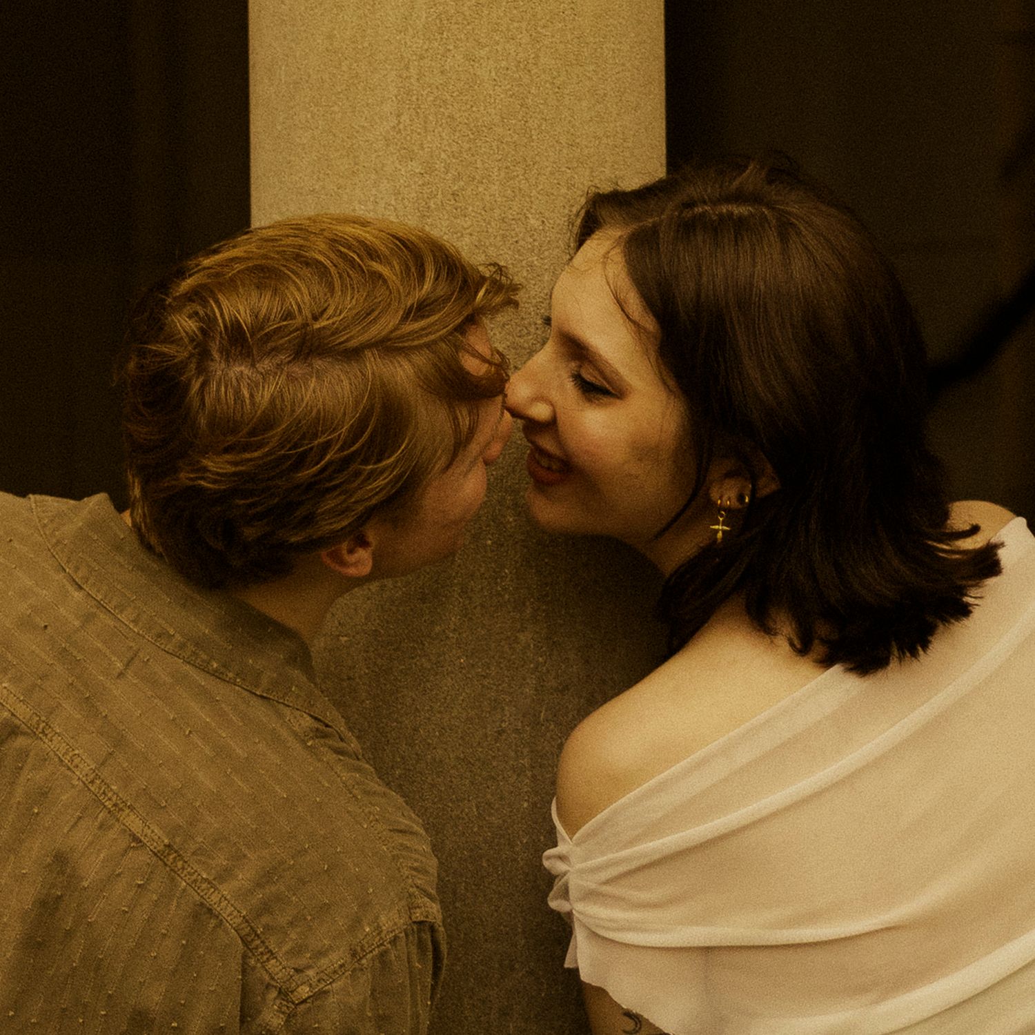 Two people in period costumes share an intimate moment, leaning close together near a stone column.