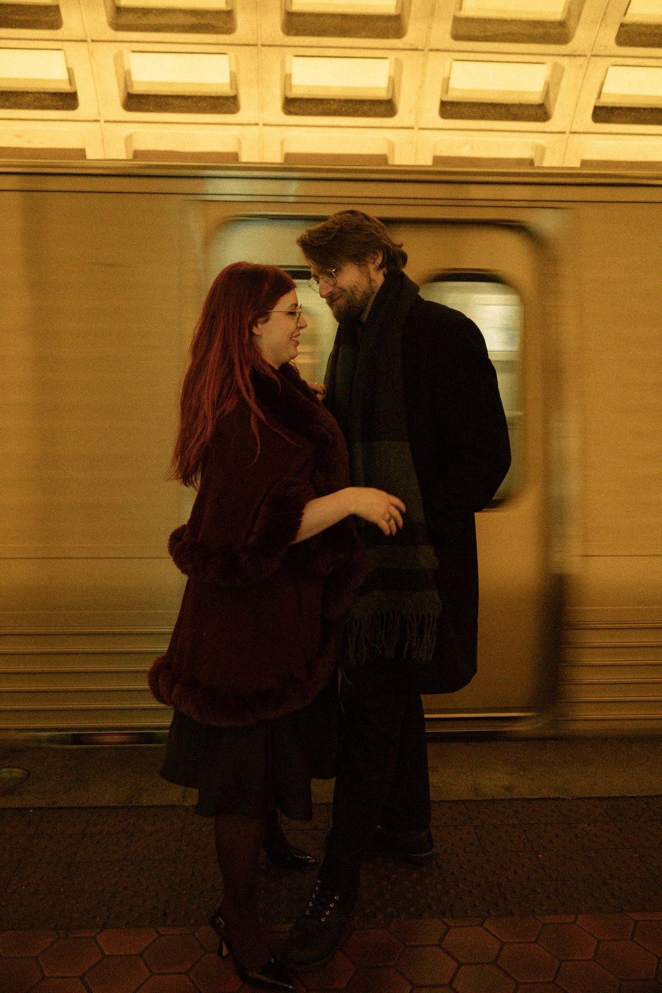 A couple in dark clothing embraces against a subway station wall in a dimly lit, sepia-toned scene.