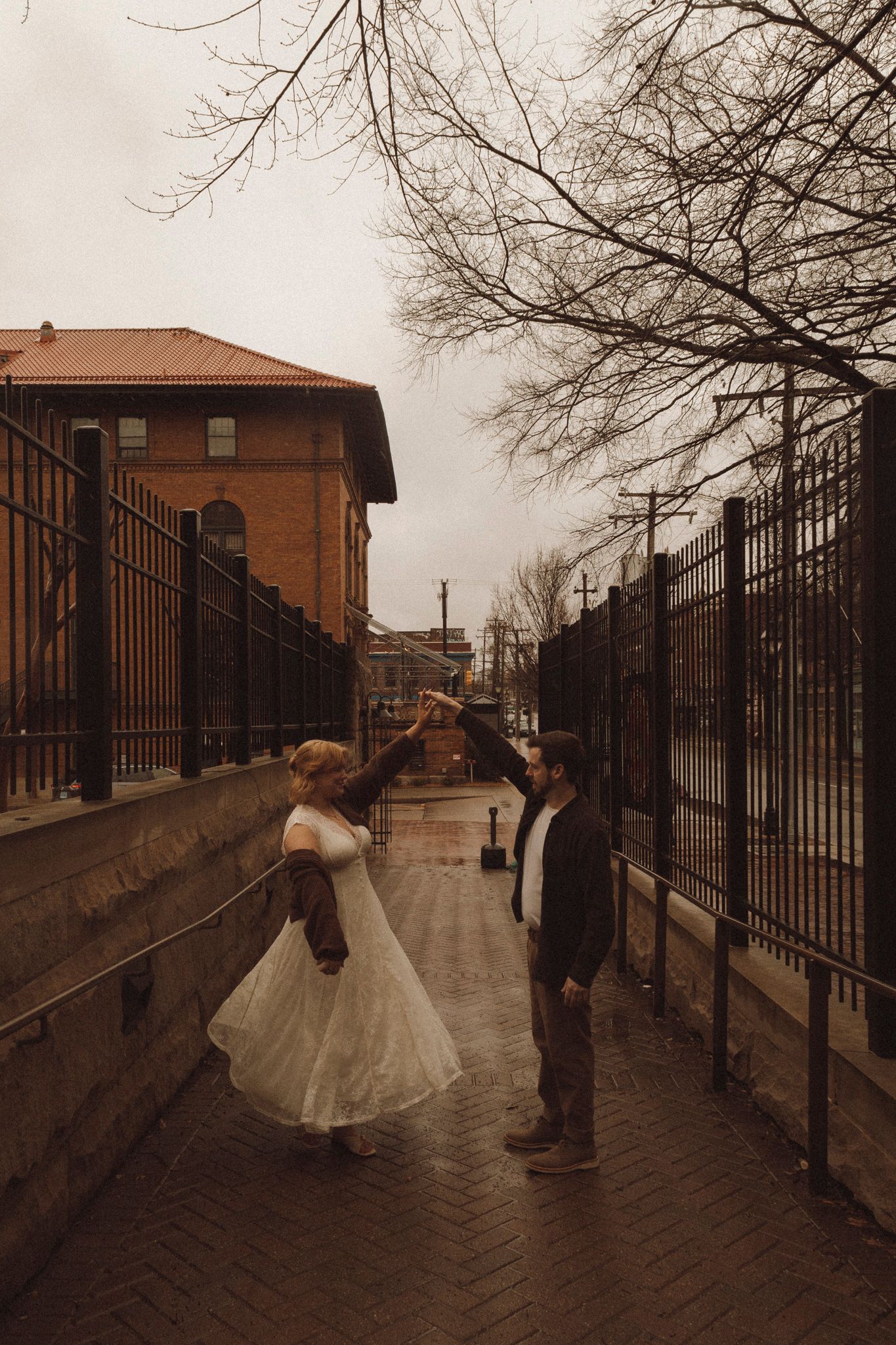 A couple in formal attire dances together on a brick walkway with buildings and bare trees in the background.