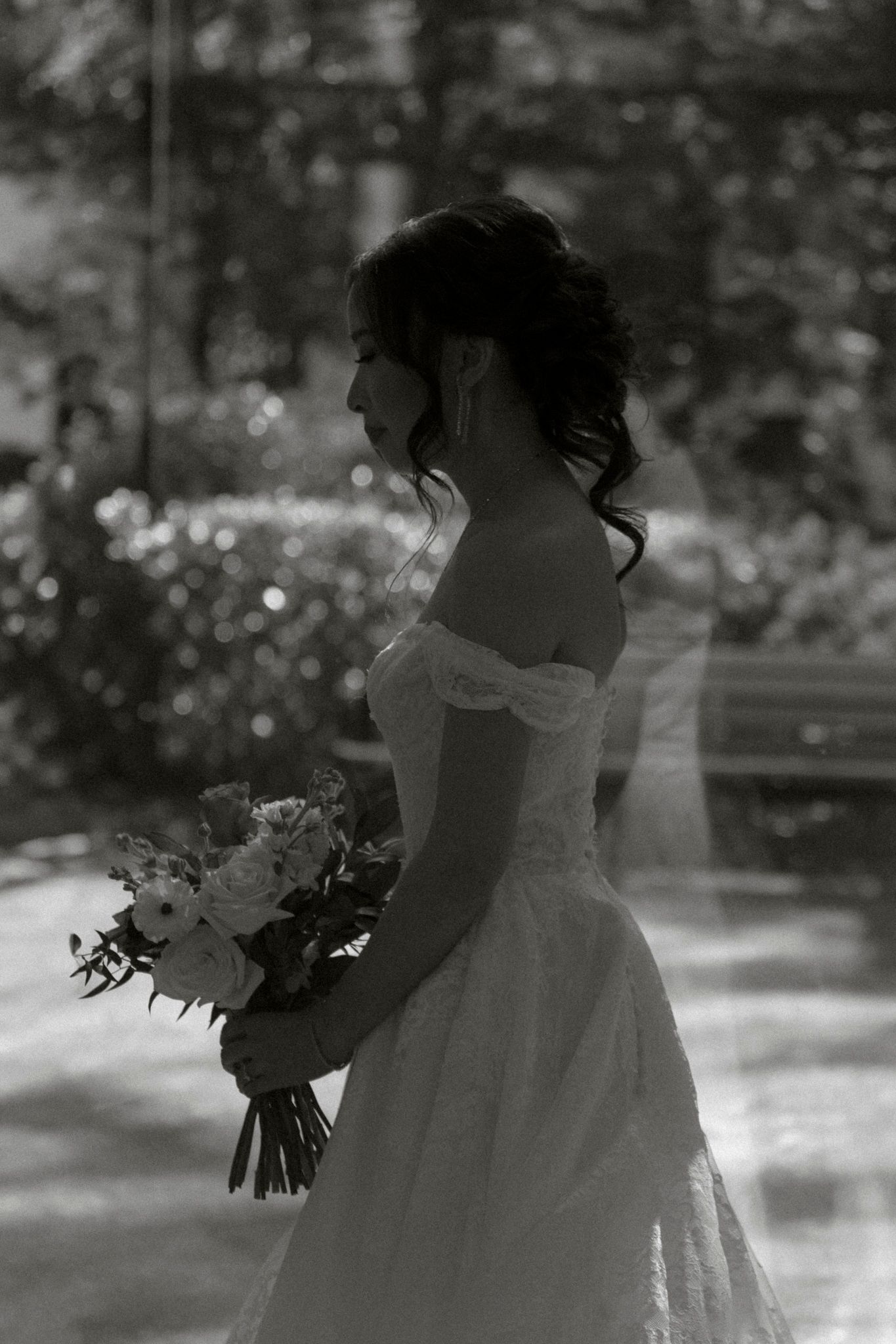 A black and white photo shows a bride in an off-shoulder wedding dress holding a floral bouquet in dappled sunlight.