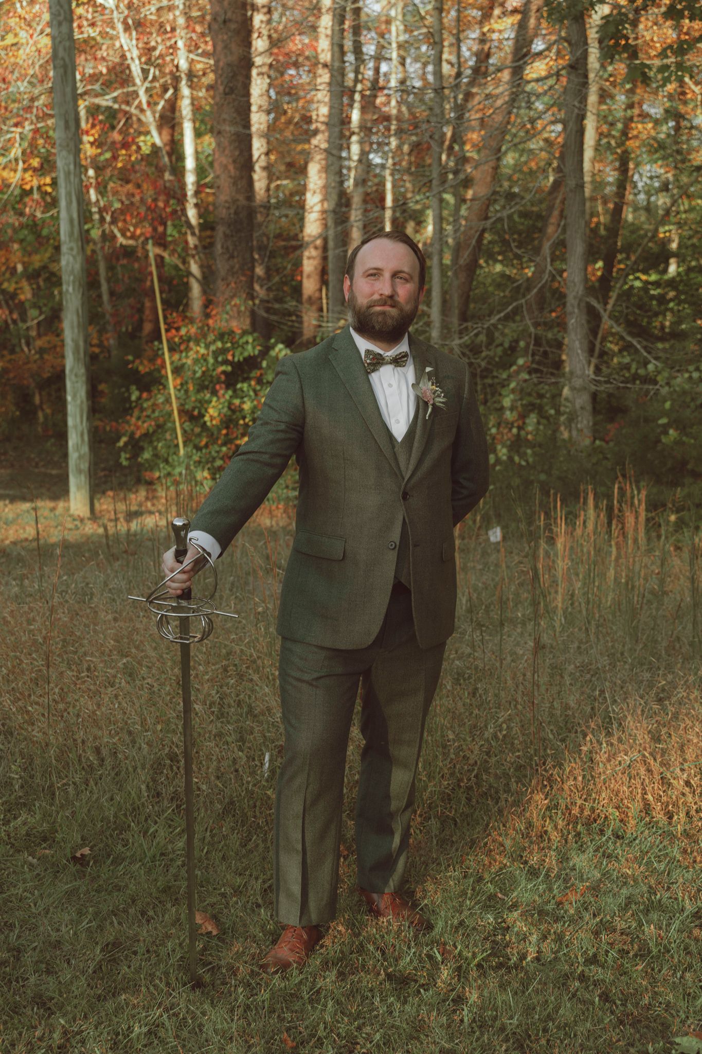 A person in a green suit stands in an autumn forest holding a cane while wearing a bowtie and boutonniere.