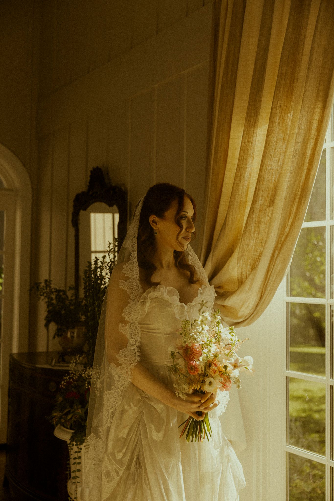 Bride in lace wedding dress and veil holds floral bouquet by sunlit window with golden curtains.