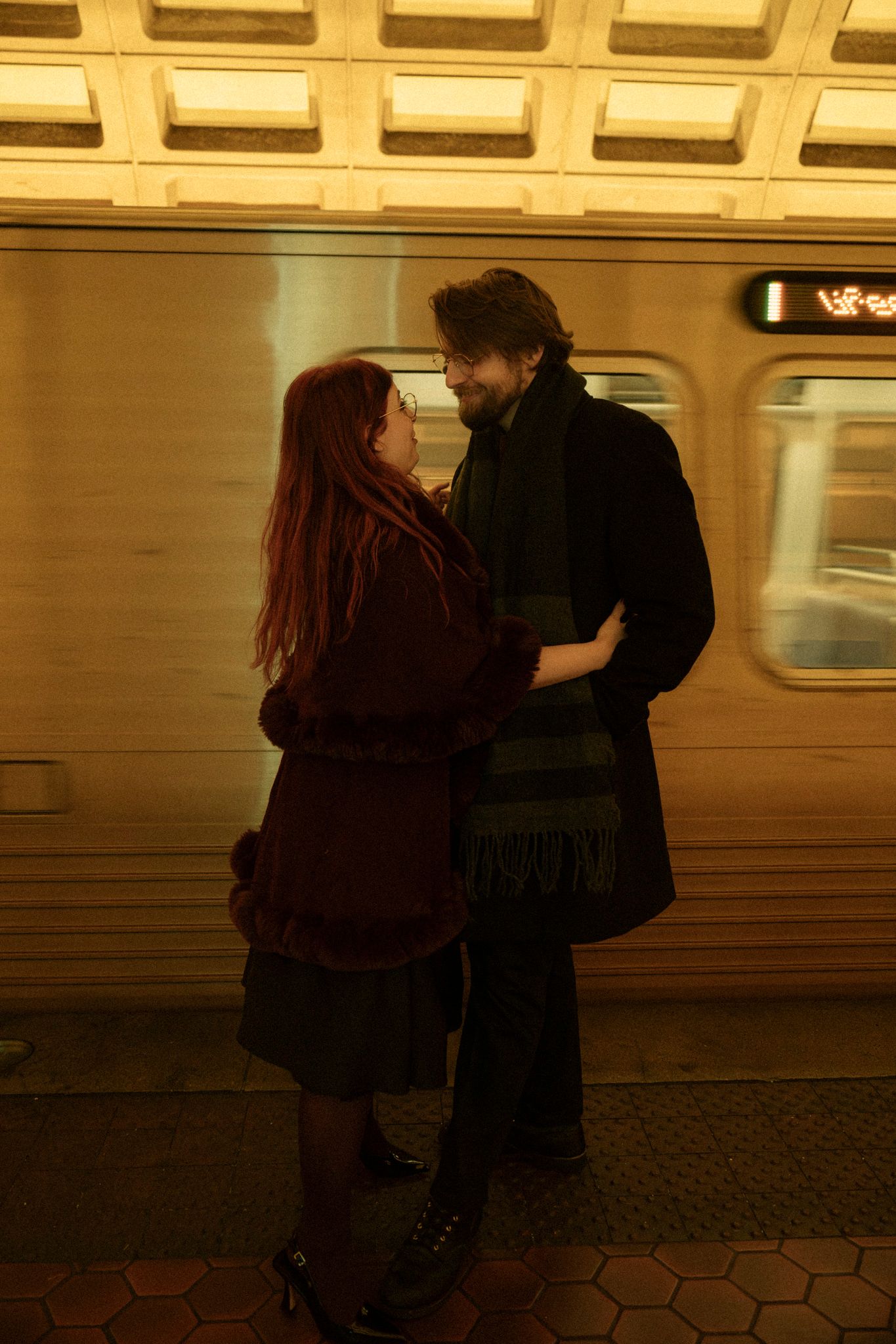A couple embraces on a subway platform as a train passes by, creating a romantic urban scene in warm lighting.