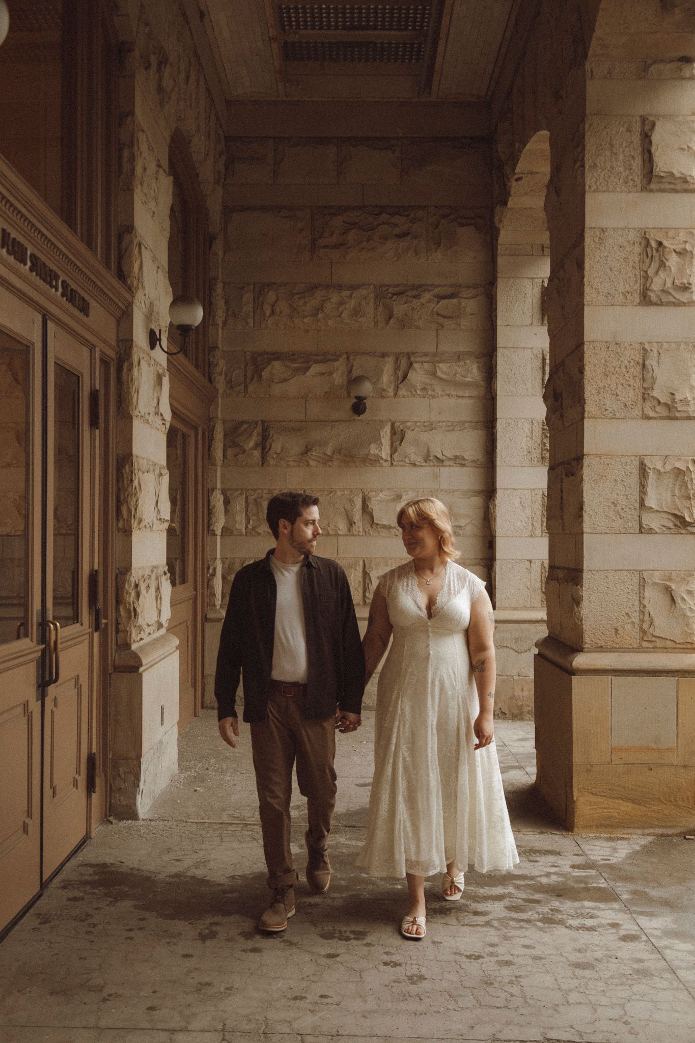 A couple walks through a rustic stone hallway in wedding attire creating a romantic vintage atmosphere.