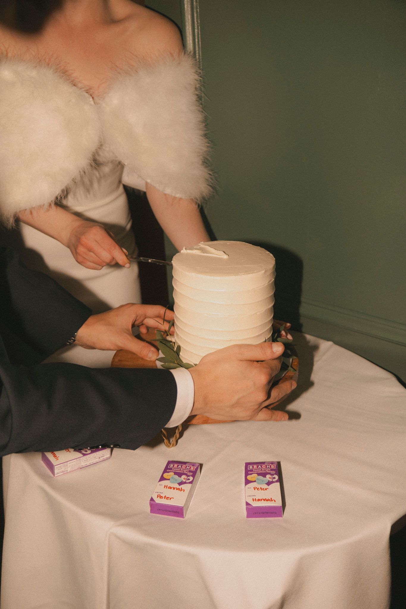 Bride and groom cutting a minimalist white tiered wedding cake on a white tablecloth table with small purple boxes nearby.