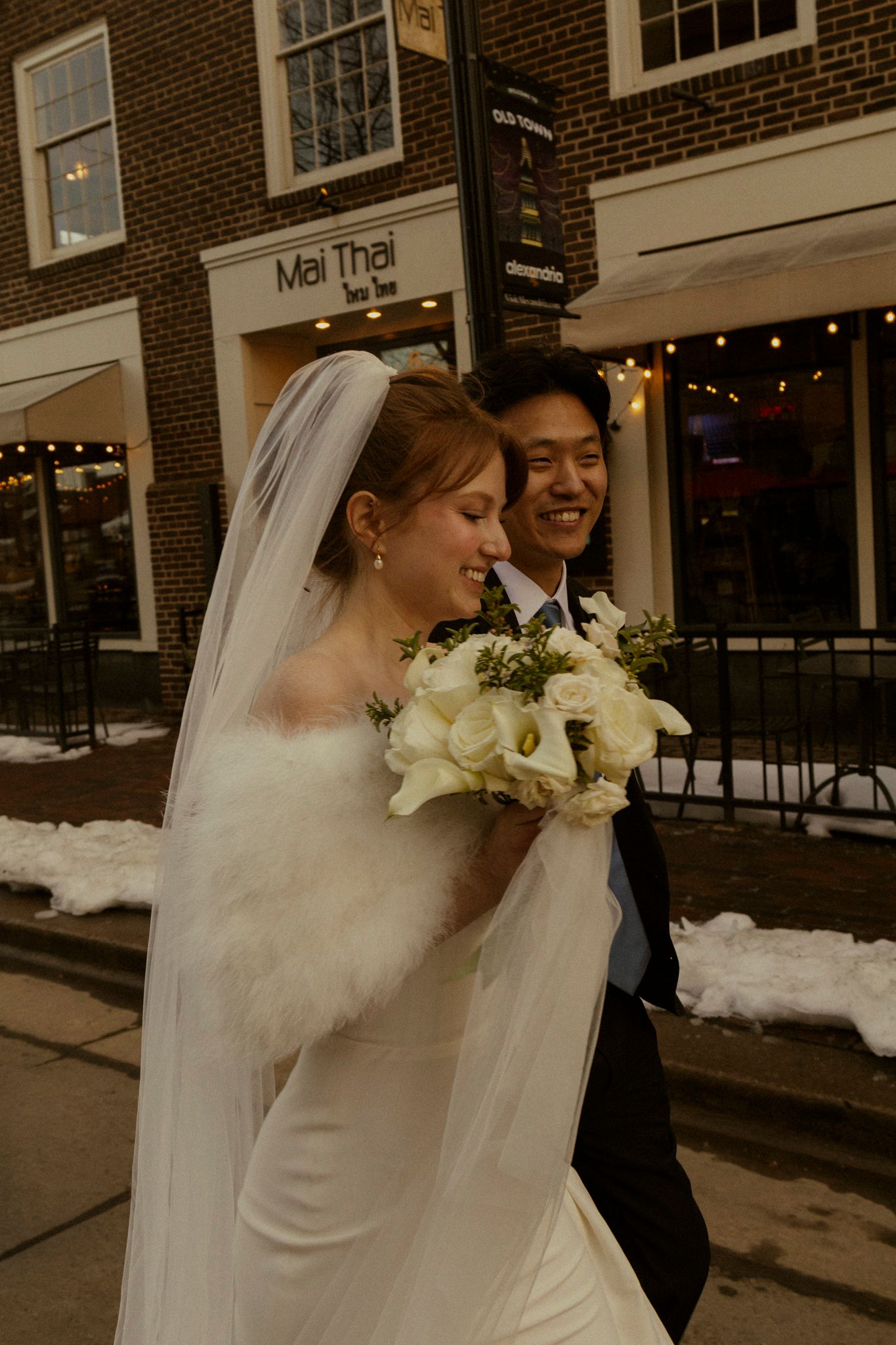 Smiling bride in white gown with fur wrap and groom walk together outside in snowy urban setting.
